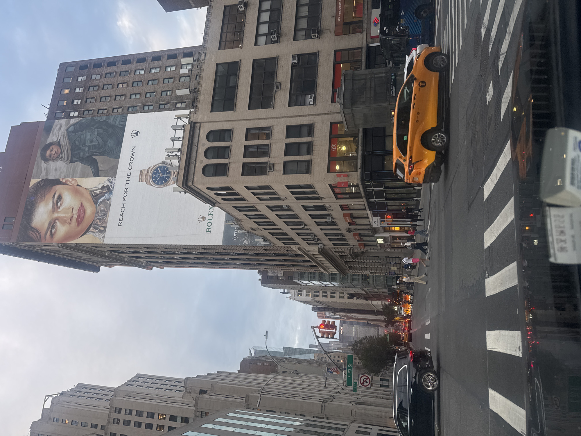 The image shows a street scene in New York City, likely Manhattan, given the urban density and architecture. The perspective is from inside a vehicle, looking forward and slightly upward.
In the foreground, a car's dashboard is visible, with some paperwork or a sticker on it. The road ahead is paved, with clear white crosswalk markings.
Dominating the middle ground are several vehicles: a black SUV on the left, an orange taxi in the center, and a blue bus on the right. Pedestrians are crossing the street at a crosswalk in the center of the image. Traffic lights are visible, with one showing red. Street signs, including "E 23 St", are also present.
The background features tall buildings lining the street, characteristic of New York City. A prominent feature is a large billboard on the side of a building, advertising Rolex, featuring a close-up of a woman's face and a watch. Another building displays "NYSC" signage. The sky is overcast, suggesting a cloudy day. The overall impression is of a busy urban intersection in New York City.