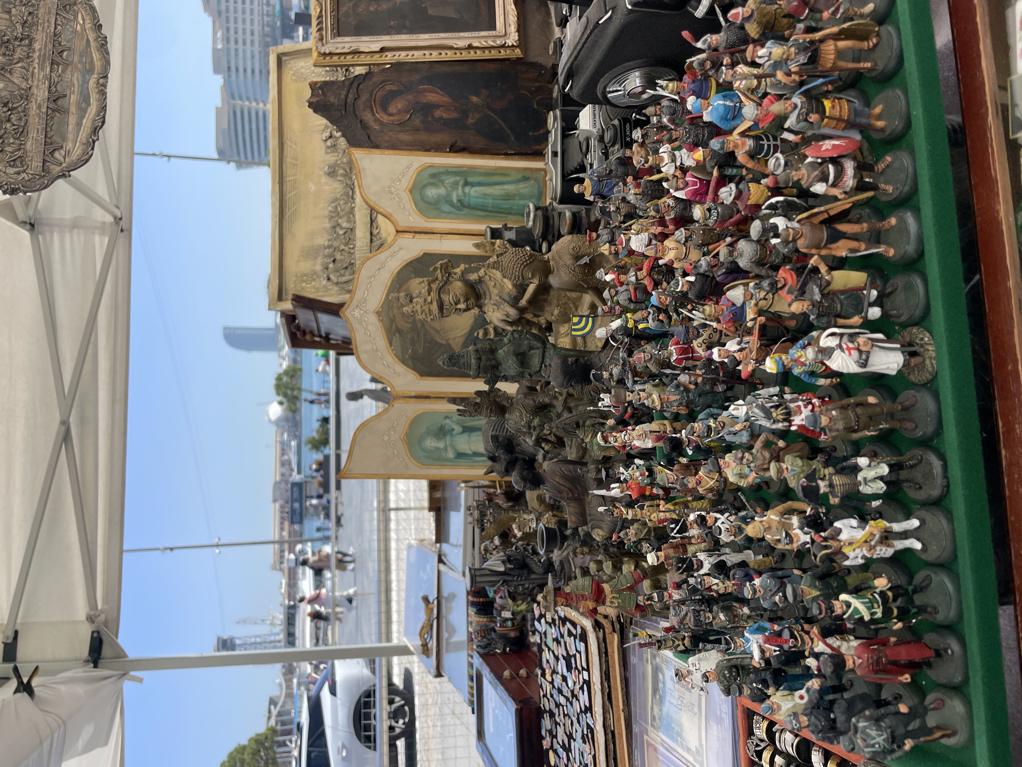 The image shows an outdoor market stall in Barcelona, Spain, likely near the port area, based on the water, boats, and buildings visible in the background. The stall displays various antique and collectible items. A large assortment of miniature figurines, mostly depicting soldiers, are arranged in rows. Behind the figurines are framed paintings, sculptures, and other objects such as an old camera. The scene is captured during daylight hours. People walk along the waterfront promenade in the background. A white car is parked nearby. The foreground of the image is cluttered with a diverse array of items for sale.