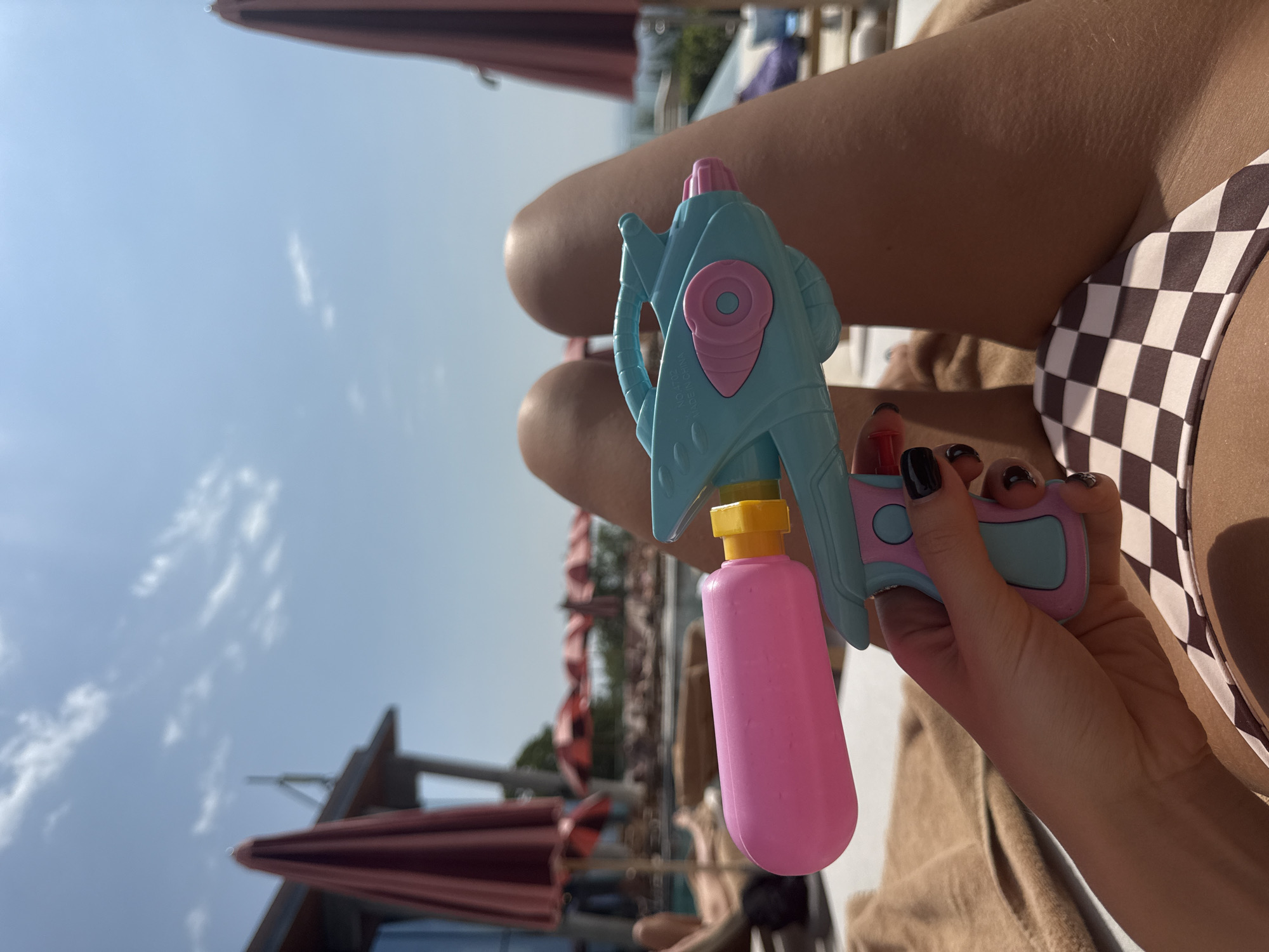 The image shows a person's hand holding a blue and pink toy water gun. The person is lying down on a towel outdoors, with their legs visible. They are wearing a checkered bikini bottom. In the background, there are lounge chairs, umbrellas, and a swimming pool, suggesting a resort or hotel setting. The sky is blue with some clouds. There is no indication of the city of Sighnaghi, Georgia in the image.