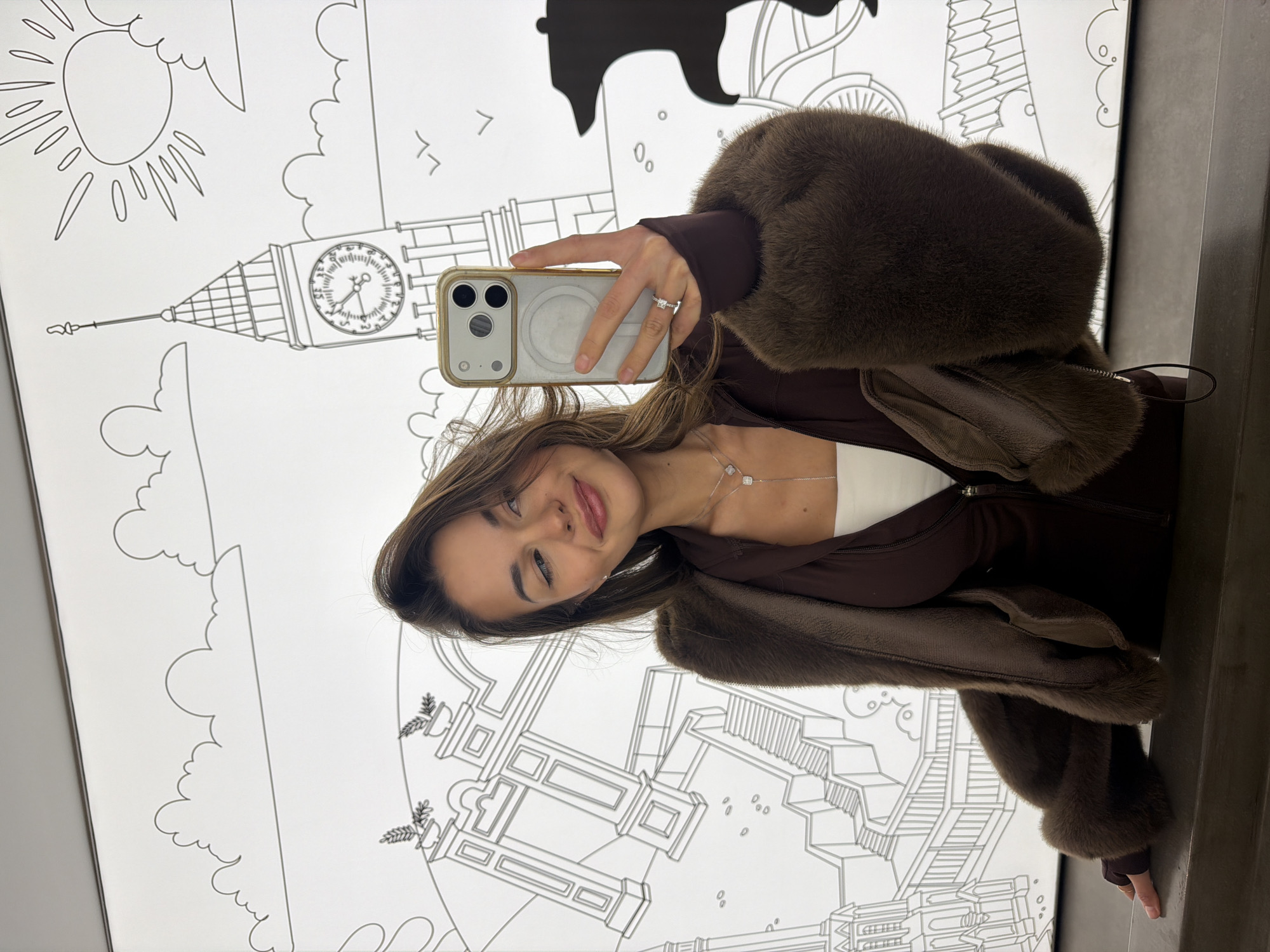 A young woman takes a selfie in front of a black-and-white mural depicting London landmarks. The mural includes the Tower of London, Big Ben, clouds, a sun, and other architectural details. She is wearing a brown fur-trimmed jacket over a brown top and a white undershirt. She is holding a white iPhone in her right hand. The woman is wearing a ring on her right hand and a necklace. The background suggests the location is indoors, possibly a studio or themed area. The mural suggests this is a tourist-focused area where visitors can take pictures with stylized depictions of global landmarks. It's unclear whether this is in Krasnoyarsk without additional context.