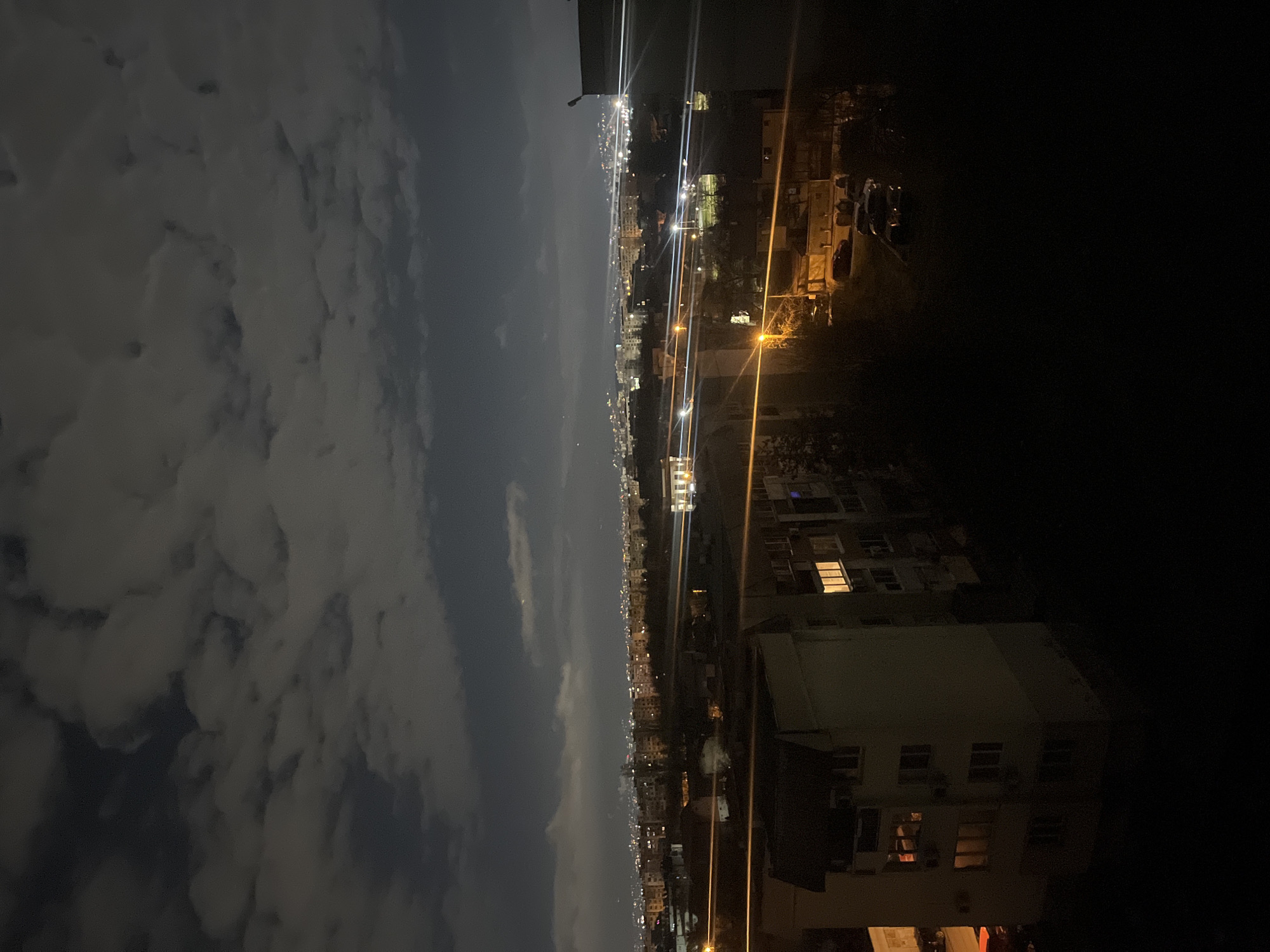 The image depicts a nighttime cityscape, most likely Sofia, Bulgaria, viewed from an elevated position. The sky is partially covered with clouds, illuminated by city lights. A line of streetlights stretches into the distance, creating streaks of light. Buildings of varying heights are visible along the cityscape, some with illuminated windows. The immediate foreground is darker, with a building and some vegetation silhouetted against the night sky. Some objects and vehicles are visible on the lower level of the image, mostly hidden in darkness.