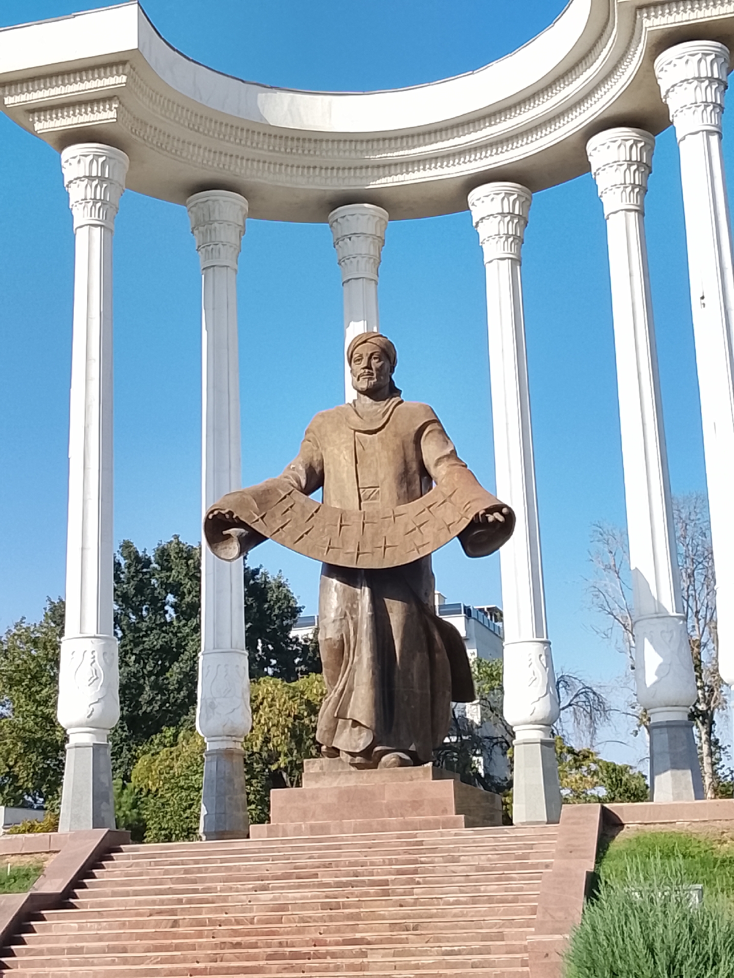 The image shows a statue of a man in traditional clothing, holding a scroll, set against a clear blue sky. The statue is located on a wide, tiered stone platform, and is surrounded by large, ornate white columns that support a curved, white canopy. Green trees are visible in the background, as well as a building. The overall scene is majestic and monumental, suggesting a historical or cultural landmark. This is the statue of Al-Fergani in Fergana City, Uzbekistan.