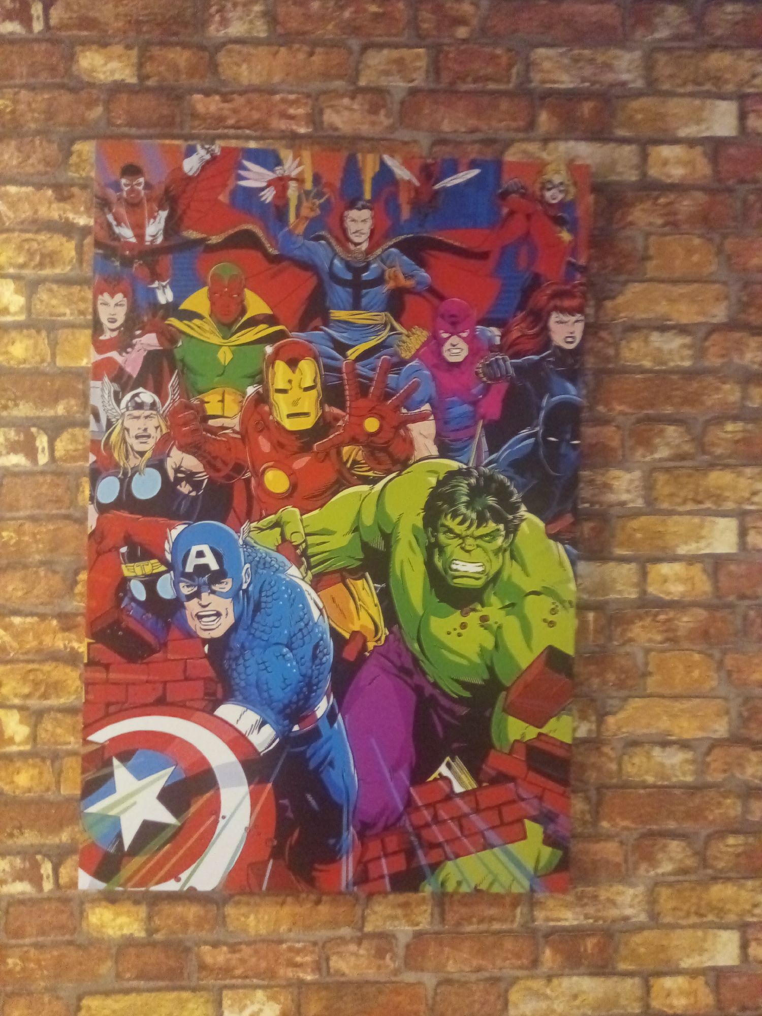 An Avengers poster is mounted on a brick wall. The poster depicts multiple superheroes in a comic book style. From top to bottom, left to right, visible characters include Falcon, Scarlet Witch, Vision, Dr. Strange, Thor, Iron Man, Captain America, Hulk, Black Widow, and Captain Marvel. The characters are arranged in a crowded composition, with many of them appearing to be in action poses. The brick wall provides a textured background for the poster. There is no indication of the specific location within Porlamar, Venezuela.