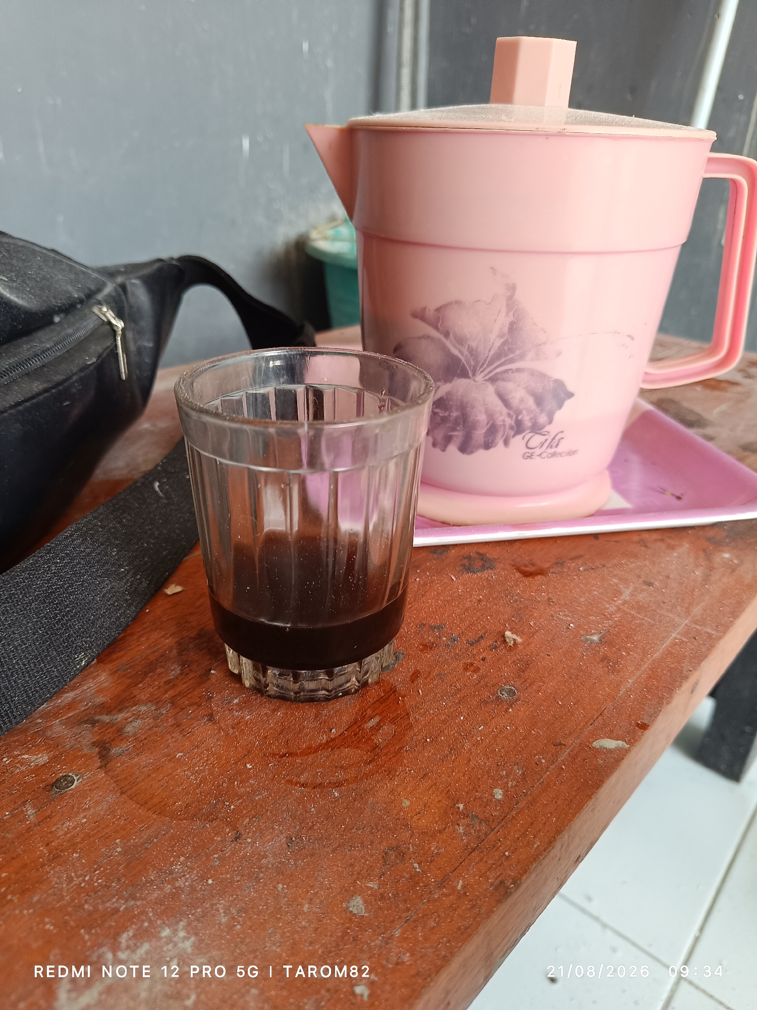 The image shows a close-up view of a wooden table with a glass of dark liquid, likely coffee, in the foreground. To the right, a pink pitcher with a lid and a floral design is visible, resting on a pink tray. A black bag with a strap is partially visible on the left. The background is blurred, but appears to be an indoor setting with a wall. A timestamp "21/08/2026 09:34" and text "REDMI NOTE 12 PRO 5G | TAROM82" are overlaid on the image, suggesting it was taken with a smartphone. The scene is consistent with a casual setting, possibly in a home or small establishment in Sruni, Indonesia.