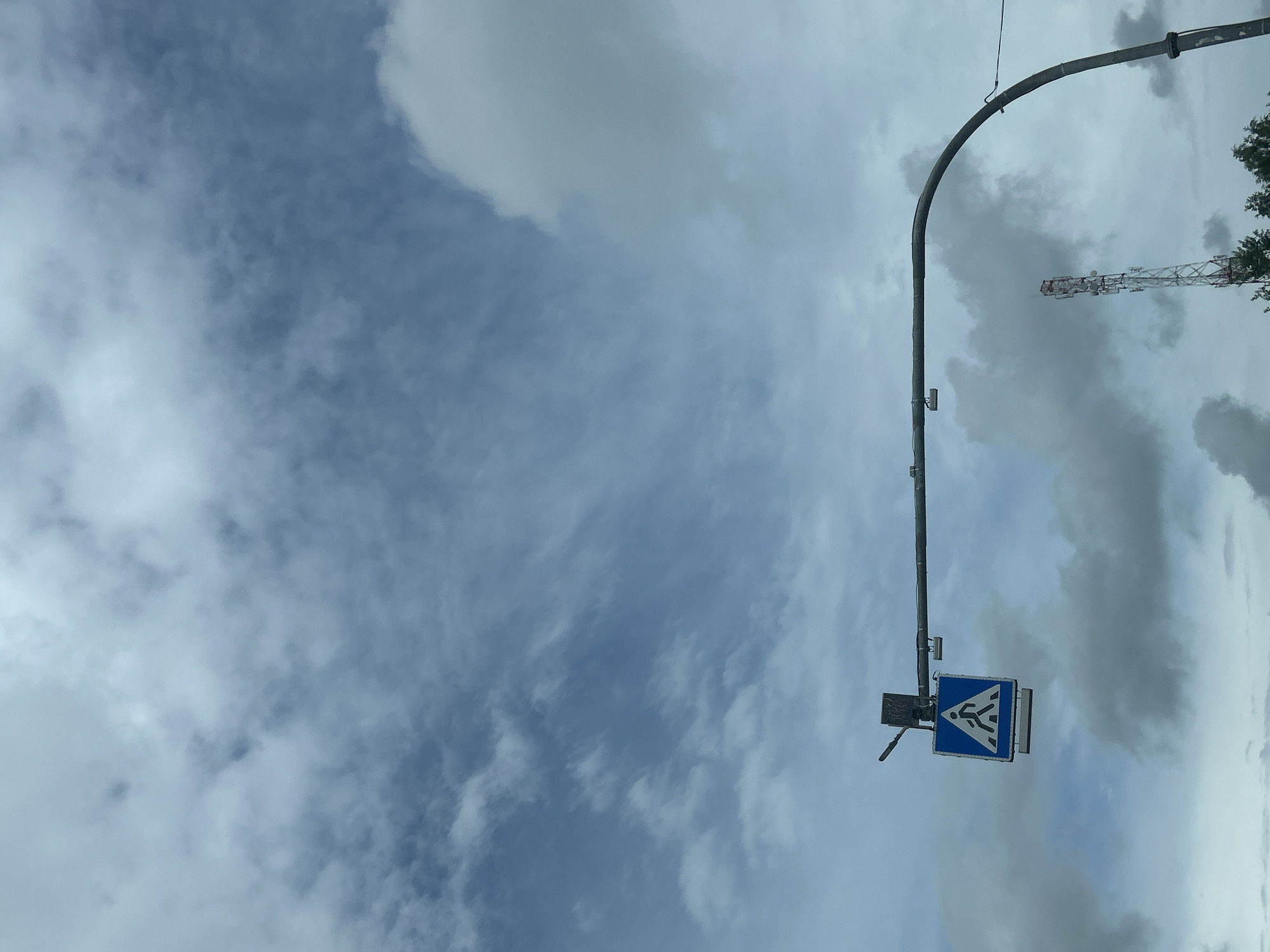 An upward-angle, medium shot captures a cloudy sky over a cityscape. Dominating the left side of the frame is a pedestrian crossing sign, a blue square with a white triangle in the center, featuring a white silhouette of a person walking. The sign is mounted on a horizontal pole, which curves downwards on the right. A tall, lattice telecommunications tower with red and white accents is visible in the background on the right, partially obscured by trees. The environment appears to be a public road area within the Плешчаніцкі сельскі Савет, Belarus. The sky is filled with a mixture of white and gray clouds, suggesting an overcast day.
