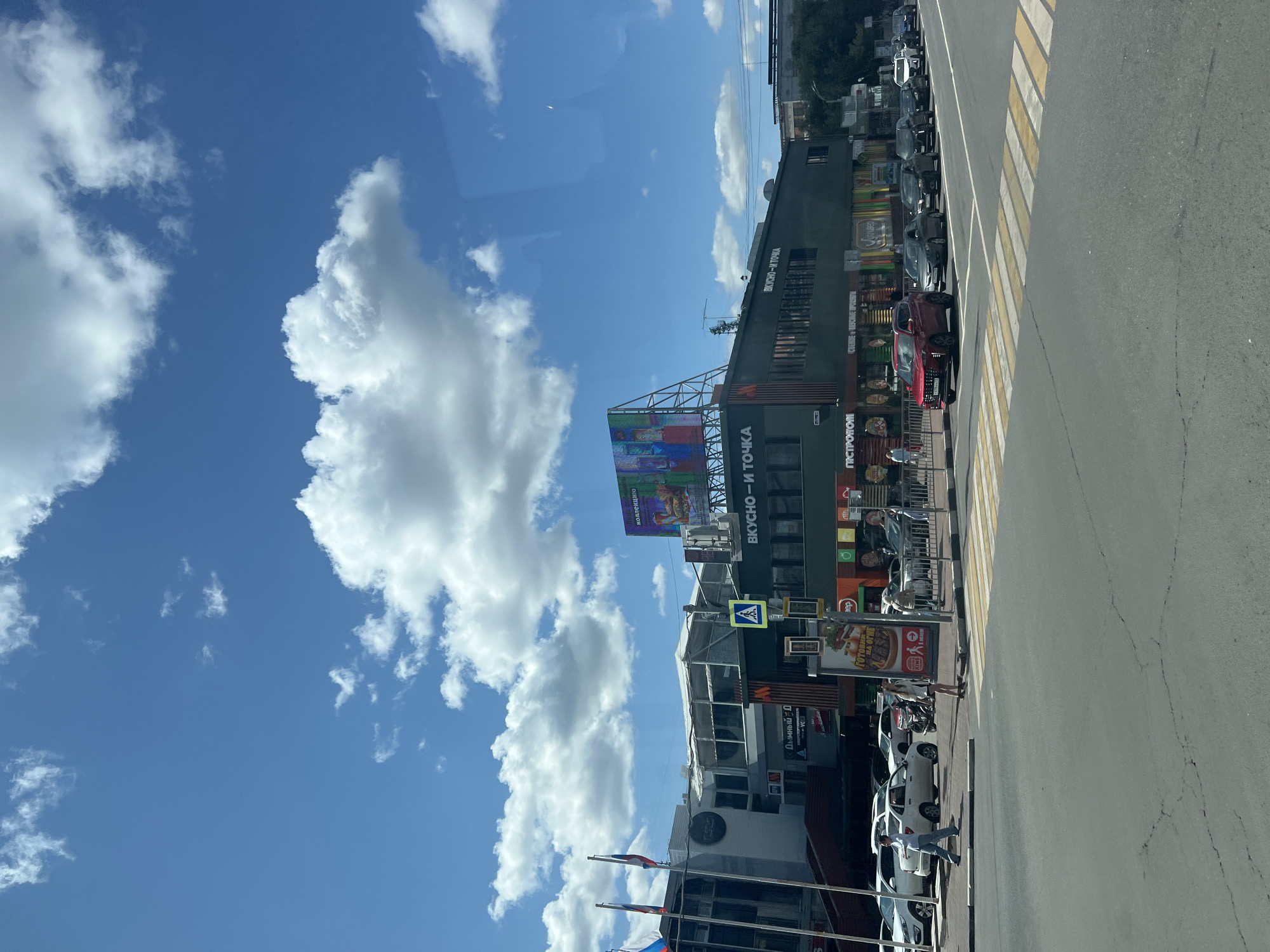 A street view in the city of Ulyanovsk, Russia, shows a busy intersection on a sunny day with a bright blue sky and scattered white clouds. On the right side of the image, a modern, dark-green building houses businesses, indicated by signage including "ВКУСНО - И ТОЧКА" (Tasty - And Point) and "ПЯТЬ РОГОВ" (Five Horns). A large digital billboard with colorful abstract patterns is mounted on a structure above the building. Several cars are lined up on the road, with a red SUV in the foreground, waiting at a crosswalk. Pedestrians are visible crossing the street and walking on the sidewalk. Russian flags fly from poles on the left side of the image, attached to a building that appears to be a government or administrative office. The scene depicts a typical urban environment with commercial activity and traffic.
