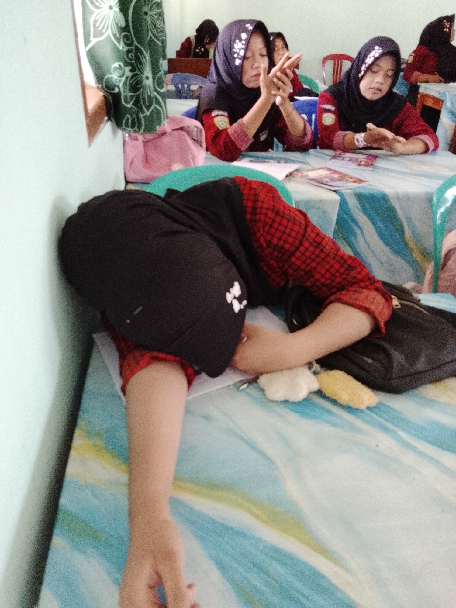 This image appears to be taken inside a classroom in Jepara, Indonesia, likely at a school. The scene depicts several young female students, all wearing hijabs and modest school uniforms, seated at tables covered with blue and white patterned cloths. The student in the foreground is lying down with her head on the table, her body angled towards the wall, wearing a black hijab and a red and black checkered shirt. Her right arm is extended on the table, and a black bag is near her. In the background, two other students are visible, also wearing hijabs and red and black shirts. One student is looking down at the table, while the other is looking forward with her hands clasped. There are various objects on the tables, including books and what appear to be decorative items. The room has light-colored walls and a window covered by a green curtain with white floral patterns. The lighting suggests an indoor setting, possibly during the daytime.