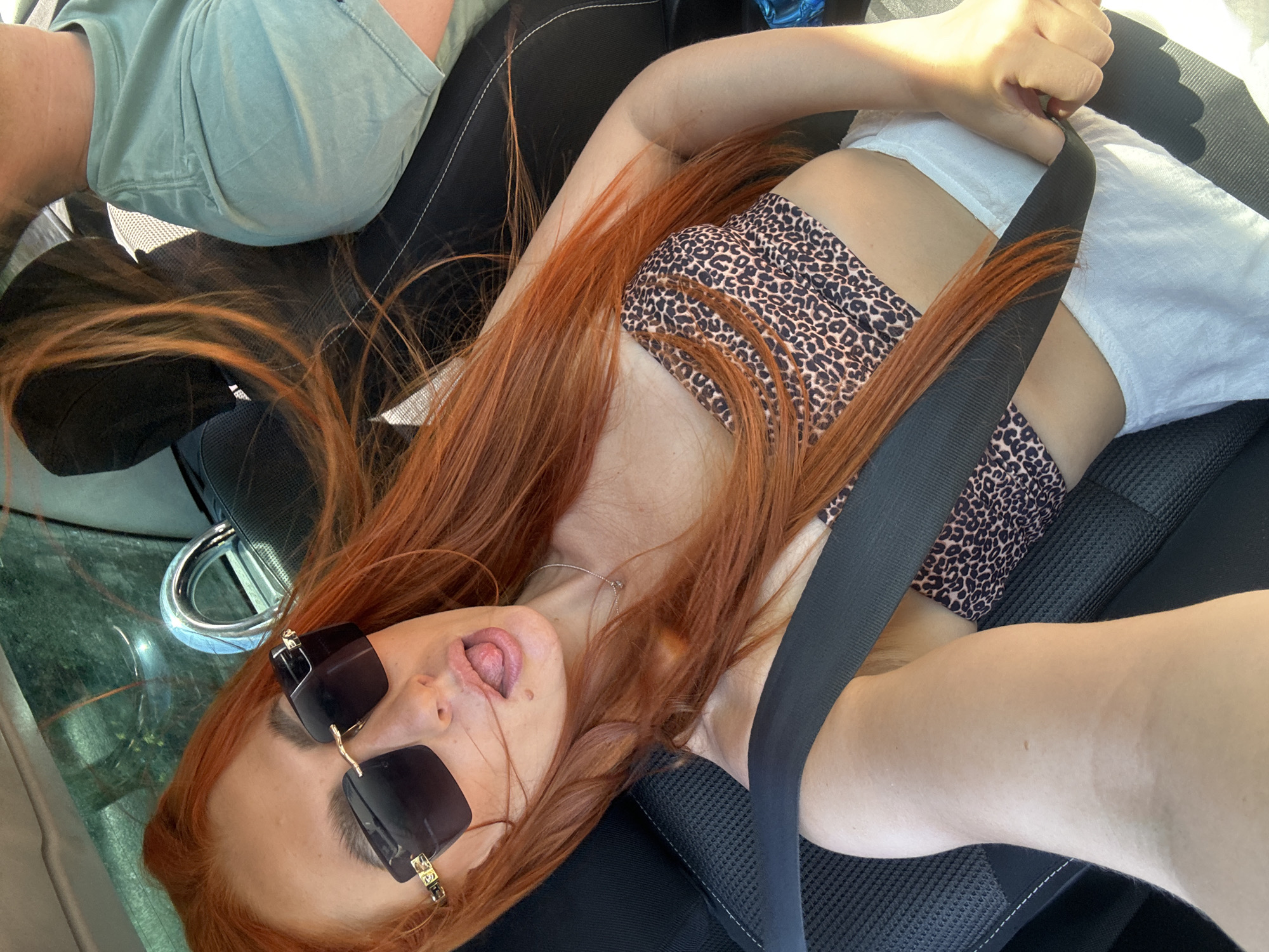 The image shows two people in a car. One person, likely a woman with long red hair, is lying down and looking at the camera with sunglasses on and her tongue sticking out. She is wearing a leopard-print bikini top and white shorts. A seatbelt is across her body. Another person, whose body is partially visible on the left of the frame, is wearing a light green t-shirt. The car appears to be parked, with a metallic loop visible on the dashboard. The scene suggests a casual outing, possibly on a sunny day. The location is identified as Golubovići, Montenegro.