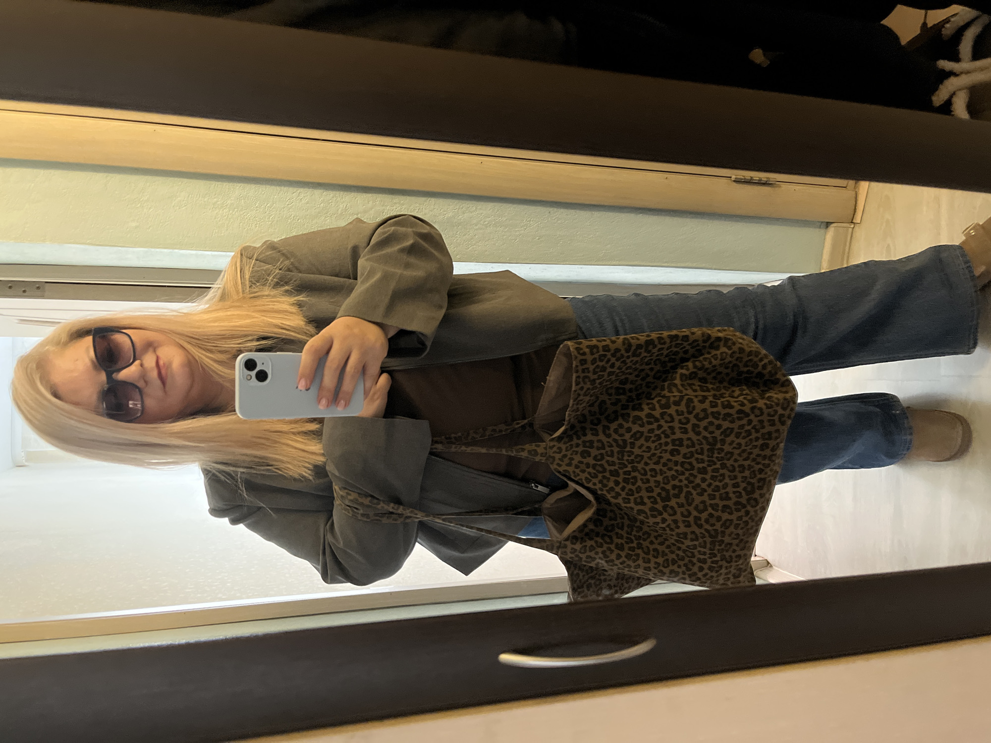 A full-body reflection of a blonde woman is captured in a mirror. The woman is wearing sunglasses, a brown top, a gray blazer, blue jeans, and tan shoes. She is holding a light blue cell phone up to take the photo. A leopard print bag hangs from her shoulder. The background shows a light green wall and dark brown trim. It is not possible to determine if the location is in Izhevsk, Russia.