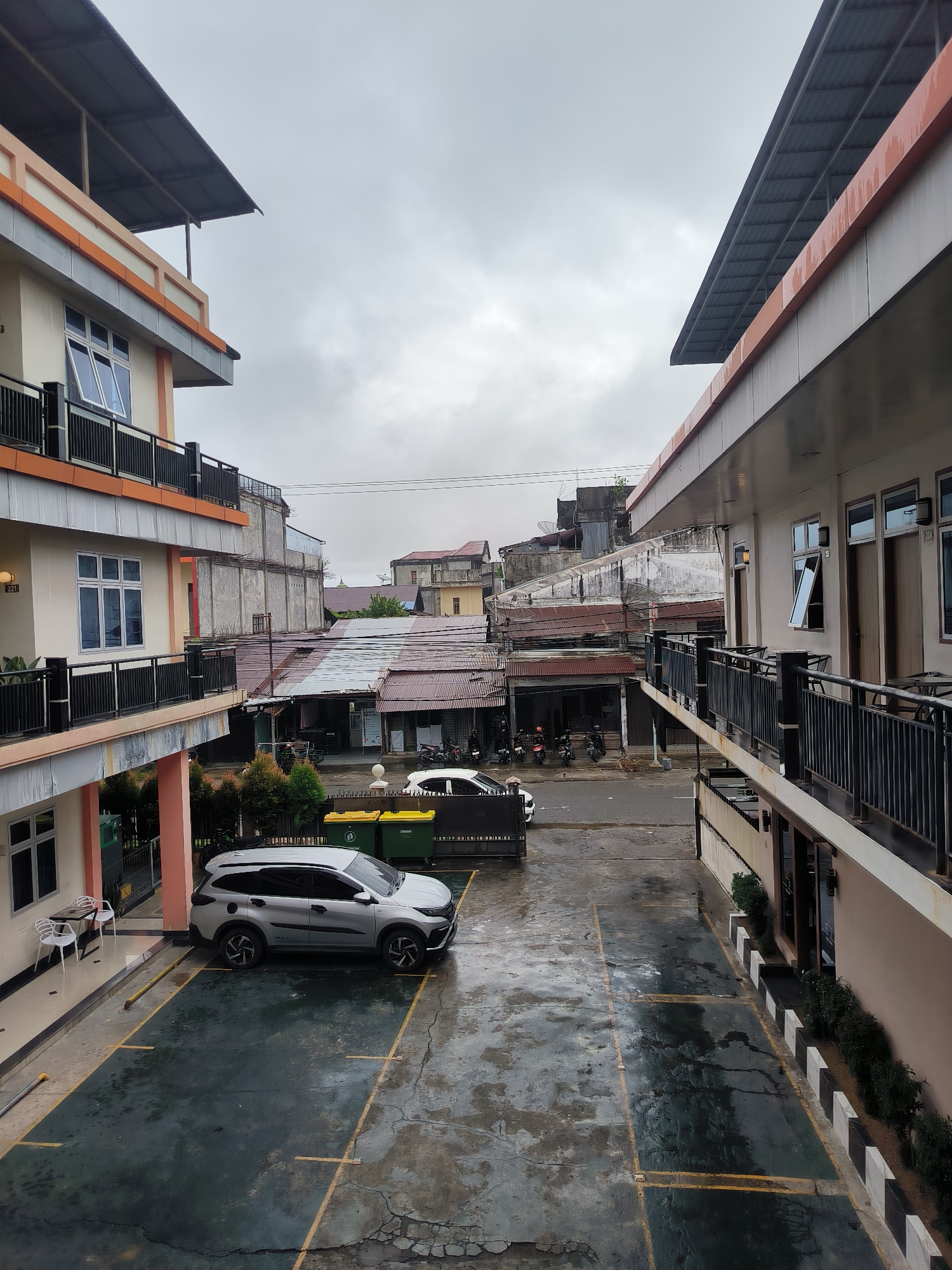 This image depicts a view from an upper floor of a building in Meulaboh, Indonesia, looking out over a street and adjacent buildings. The foreground features a wet asphalt parking lot with yellow parking lines. A silver SUV is parked in one of the spots. To the left, part of the building's exterior is visible, including a balcony with black railings and windows. Two white chairs and a small table are placed on a paved area near the building.
In the middle ground, the street is wet, suggesting recent rain. Several motorcycles are parked along the side of the street. Adjacent to the street are buildings with corrugated metal roofs, some appearing older and weathered. A few windows and doors are visible on these structures. Two large green and yellow trash bins are situated near the edge of the parking lot.
The background shows more buildings, some of which have satellite dishes. The sky is overcast, with a grayish hue. The overall impression is that of a moderately urban environment, possibly a residential or commercial area, under cloudy weather conditions. There are no people clearly visible in the image.