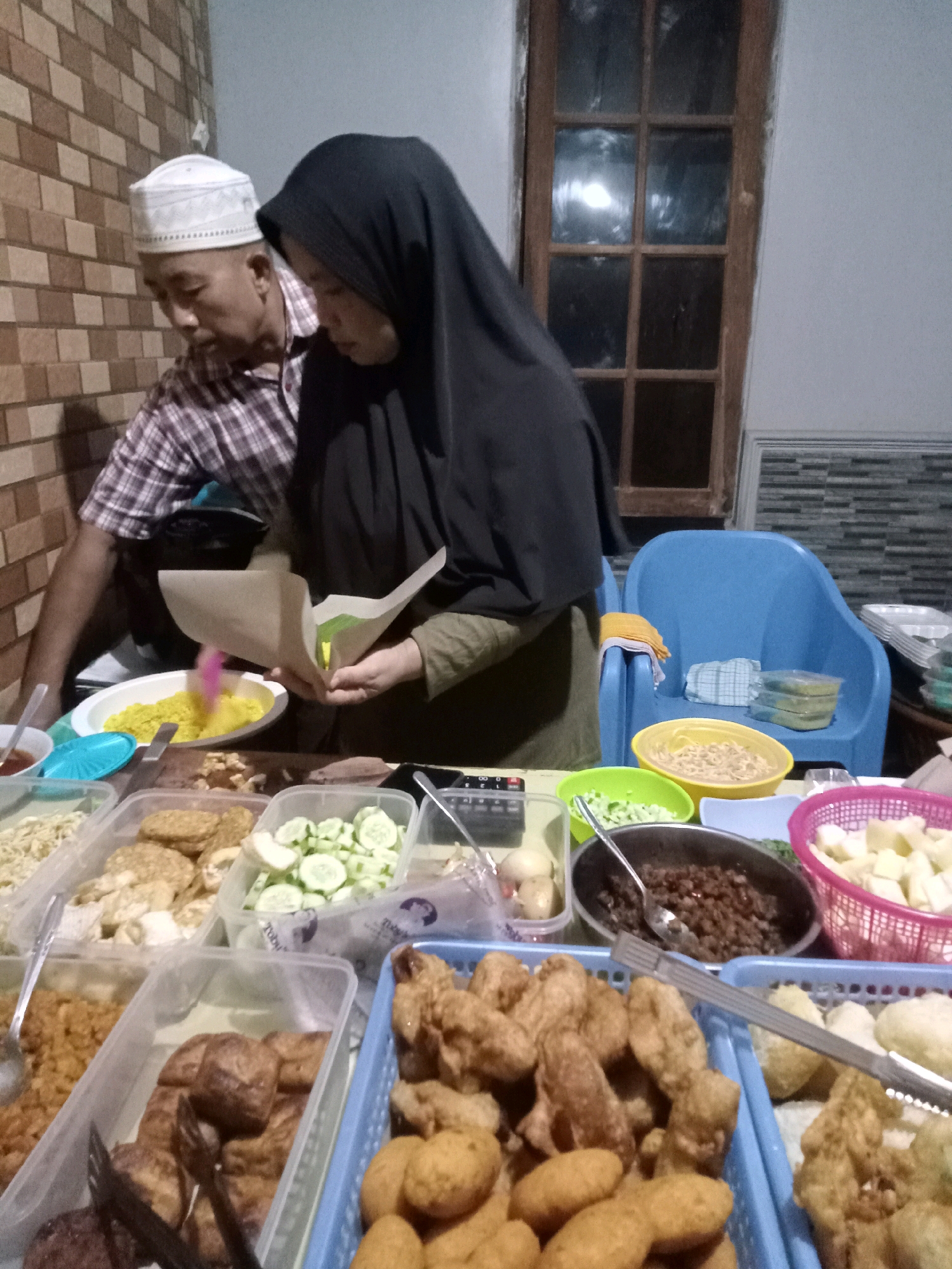 In Cirebon, Indonesia, a man wearing a white peci and a plaid shirt stands beside a woman in a black hijab, both engaged in preparing food. The woman is seen holding a paper cone, likely for serving, and a pink-handled utensil is placed in a bowl of yellow rice. A variety of food items are displayed in plastic containers and baskets, including what appears to be fried tempeh, sliced cucumbers, noodles, and possibly fried chicken. A blue plastic chair is visible in the background, along with a window with wooden panes. The scene suggests a street food stall or a home-based food preparation area, indicated by the casual setting and the array of food items.