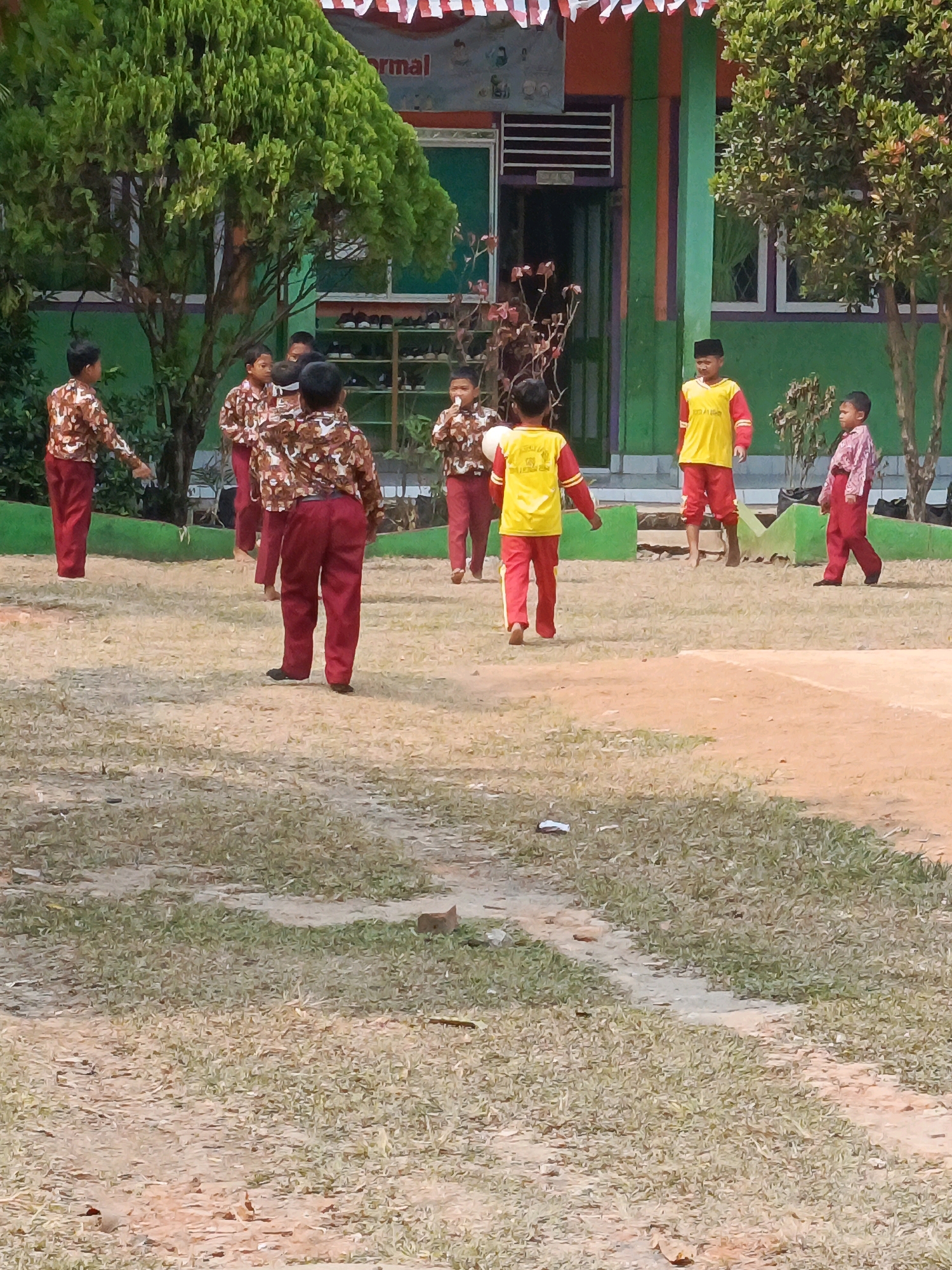 This is an outdoor photograph taken during daylight at what appears to be a school in Megang Sakti IV, Indonesia. The scene depicts a group of young boys in school uniforms engaged in some activity on a grassy and dirt courtyard. The building in the background is painted in green and orange with decorative colorful vertical stripes. In front of the building, there is a row of green planters. The courtyard is mostly covered in dry grass and patches of dirt.
There are at least seven boys visible in the frame. Several boys are wearing patterned shirts with brown and white camouflage-like designs and red pants. One boy in the center foreground is wearing a yellow shirt with red sleeves and red pants, and a black cap. Another boy to the right is wearing a pink patterned shirt and red pants. The boys are scattered across the courtyard, with some appearing to be moving or interacting. One boy on the left is pointing forward with his left hand. The overall atmosphere seems to be one of casual play or a school activity.