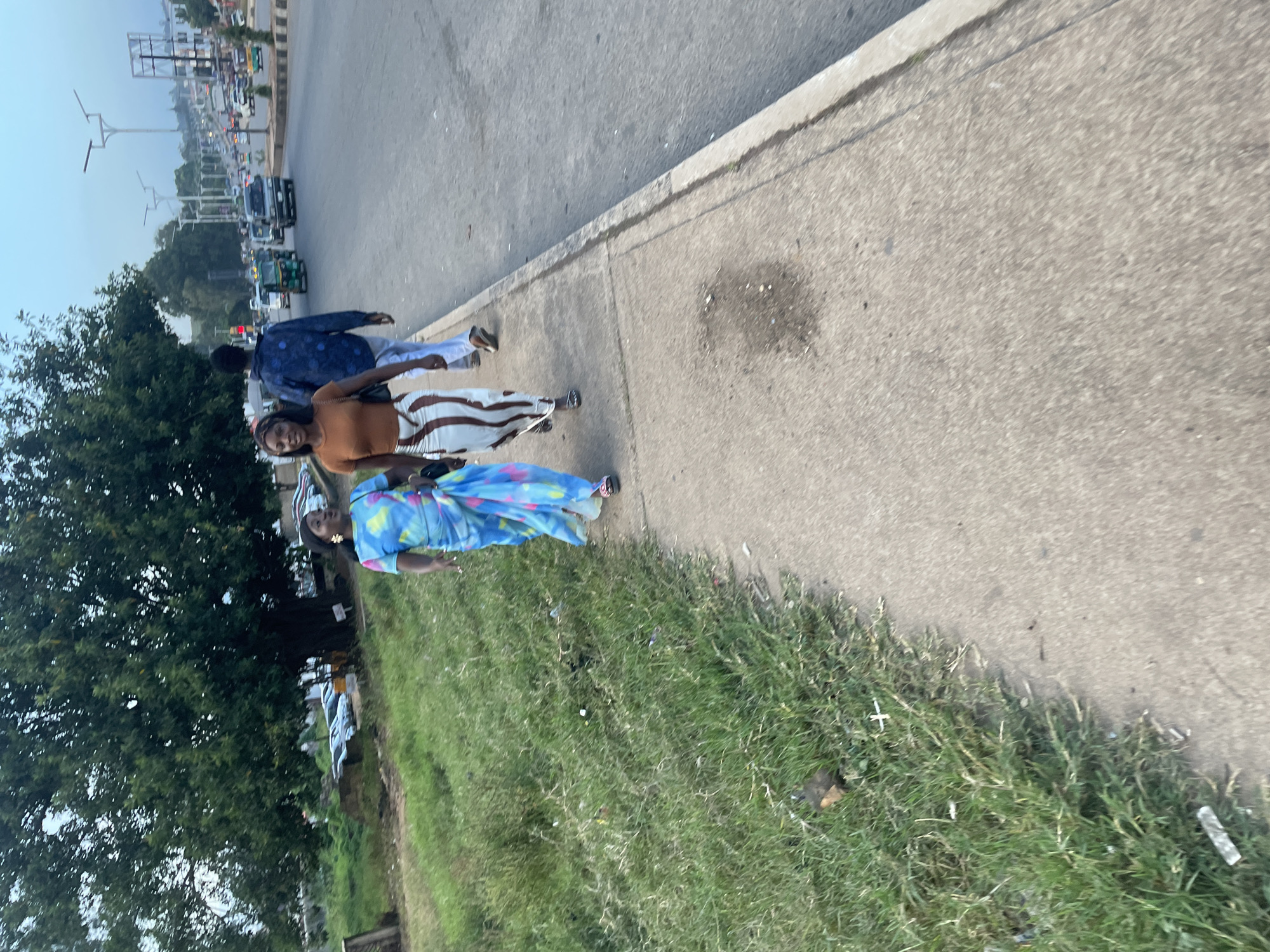 The image captures a sidewalk scene in Gura Top, Nigeria. In the foreground, two women are walking on a paved sidewalk. The woman on the left wears a brightly colored tie-dye dress, and the woman on the right wears a brown top and a striped skirt. Behind them, a man in a blue patterned shirt and white pants is also walking. The sidewalk is adjacent to a grassy area with some debris. Across the street, a busy road is visible with various vehicles, including cars and a three-wheeled auto-rickshaw, suggesting an urban environment. The background shows more of the city, with buildings and streetlights under an overcast sky.