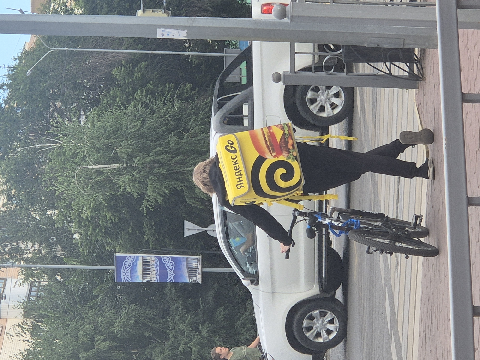 In Kokshetau, Kazakhstan, a person carrying a yellow insulated delivery backpack with a burger image and "Яндекс Go" branding is walking a bicycle across a pedestrian crossing. The person is wearing black pants and dark shoes. Behind them, a white pickup truck is stopped at an intersection, and a male in a green shirt is standing to the left of the truck. A tall street lamp with a blue banner featuring architectural elements and ornate patterns stands near the intersection, surrounded by green trees and buildings. The sky is visible overhead.