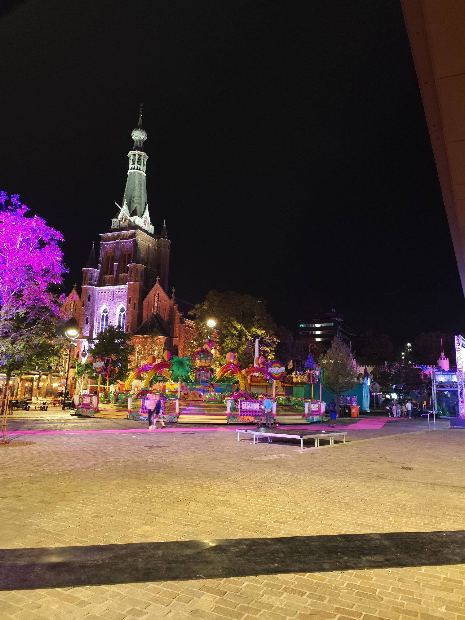 This is a night-time, long-exposure photograph of an outdoor fair or carnival in the city of Tilburg, Netherlands, specifically in the central area, likely near the Heuvelplein or Piusplein, given the presence of a prominent church tower.
In the background, a large brick church with a tall, pointed spire is illuminated by purple and white lights, highlighting its architectural features. To the left of the church, a tree is bathed in vibrant pink light. In front of the church, various carnival rides and attractions are visible, brightly lit with colorful lights, including a spinning ride with vibrant rainbow-colored elements and what appears to be a smaller children's ride. Some of these rides feature playful, cartoonish designs.
In the mid-ground, several people are walking across a paved area. Two individuals are in motion, appearing as streaks of light due to the long exposure. Other people are standing and observing the attractions, some gathered in small groups. There are also tables and chairs from outdoor cafes or restaurants visible on the left side.
The foreground consists of a paved plaza made of light-colored bricks, illuminated by a warm, yellow light, creating a reflective surface. A dark, rectangular shadow is cast across the lower portion of the plaza. The sky is dark and clear, typical of a night scene, with a few faint stars visible. The overall atmosphere is lively and festive, with the contrast between the illuminated carnival and the dark night sky.