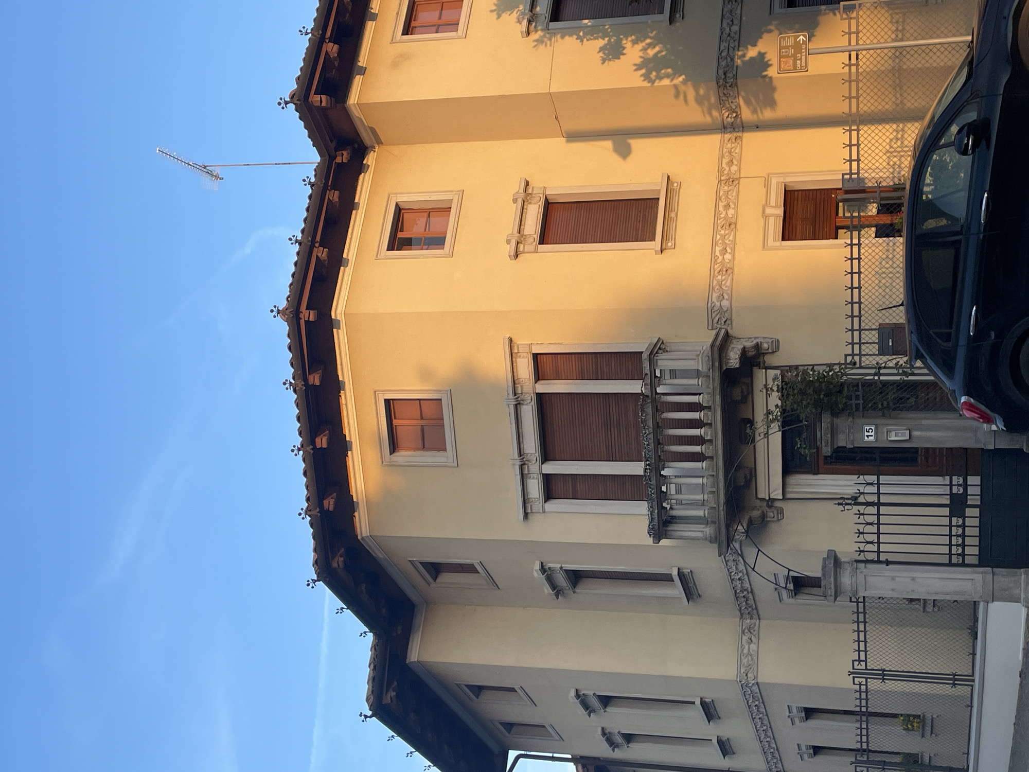 The photograph depicts the facade of a multi-story building in Case/Scjasis, Italy. The building is painted in a pale yellow hue and features windows with dark brown shutters. A wrought-iron balcony is located on the first floor. A black vehicle is parked alongside the building. A visible sign displays "Case / Scjasis". The sky is clear blue.