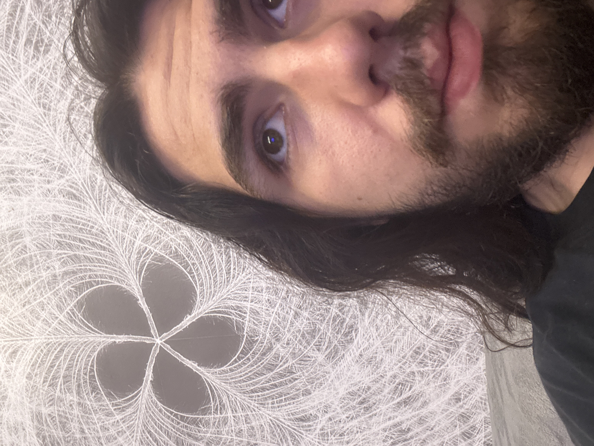 A close-up, low-angle, waist-up shot shows a man with long, dark hair and a beard. He is looking up and to his right. The man is wearing a dark t-shirt. The background is a textured wallpaper with a white, feather-like pattern that resembles a snowflake or a flower. The lighting is warm and diffused, creating soft shadows. The image appears to have been taken indoors. There is no identifiable location in the city of Unknown, Unknown.
