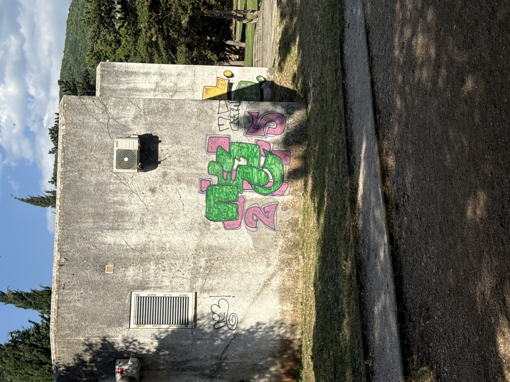 The image shows the exterior of a concrete building with graffiti. The building is light grey and appears weathered. There is a rectangular window with horizontal blinds on the left side. An air conditioning unit is mounted on the wall above and to the left of the window. Another air conditioning unit is mounted higher on the wall on the right side of the building.
The graffiti on the wall consists of stylized letters and numbers in bright colors. A prominent green tag with pink outlines is in the center of the wall. To the left of this tag, there is black graffiti that appears to be a script with a spiral and an exclamation mark. To the right, there are some yellow and green shapes, also appearing to be part of a tag.
In front of the building, there is a grassy area. Beyond the building, trees and a hillside are visible. The sky is blue with scattered clouds. The scene appears to be in an outdoor public space. The graffiti suggests an urban environment, and the style of the building and the surrounding vegetation could be consistent with locations in Budva, Montenegro, particularly in areas with older infrastructure. The light suggests it is daytime, possibly late afternoon given the shadows.