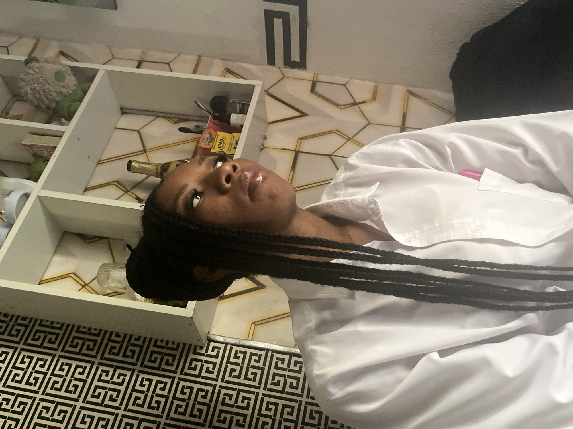 A young Black woman with long, braided hair is shown in a close-up shot, looking upwards and to her right. She is wearing a white button-down shirt. The image appears to be taken indoors, with shelves visible in the background. On the shelves, there are various objects including a green stuffed toy, books, bottles, and containers. The background wall has a geometric pattern. The floor also has a black and white geometric tile pattern. The lighting suggests it could be daytime. The woman's expression is neutral. A small pink object, possibly a pen, is tucked into the front of her shirt. There is no discernible text in the image. The context provided indicates the location is Jos, Nigeria.