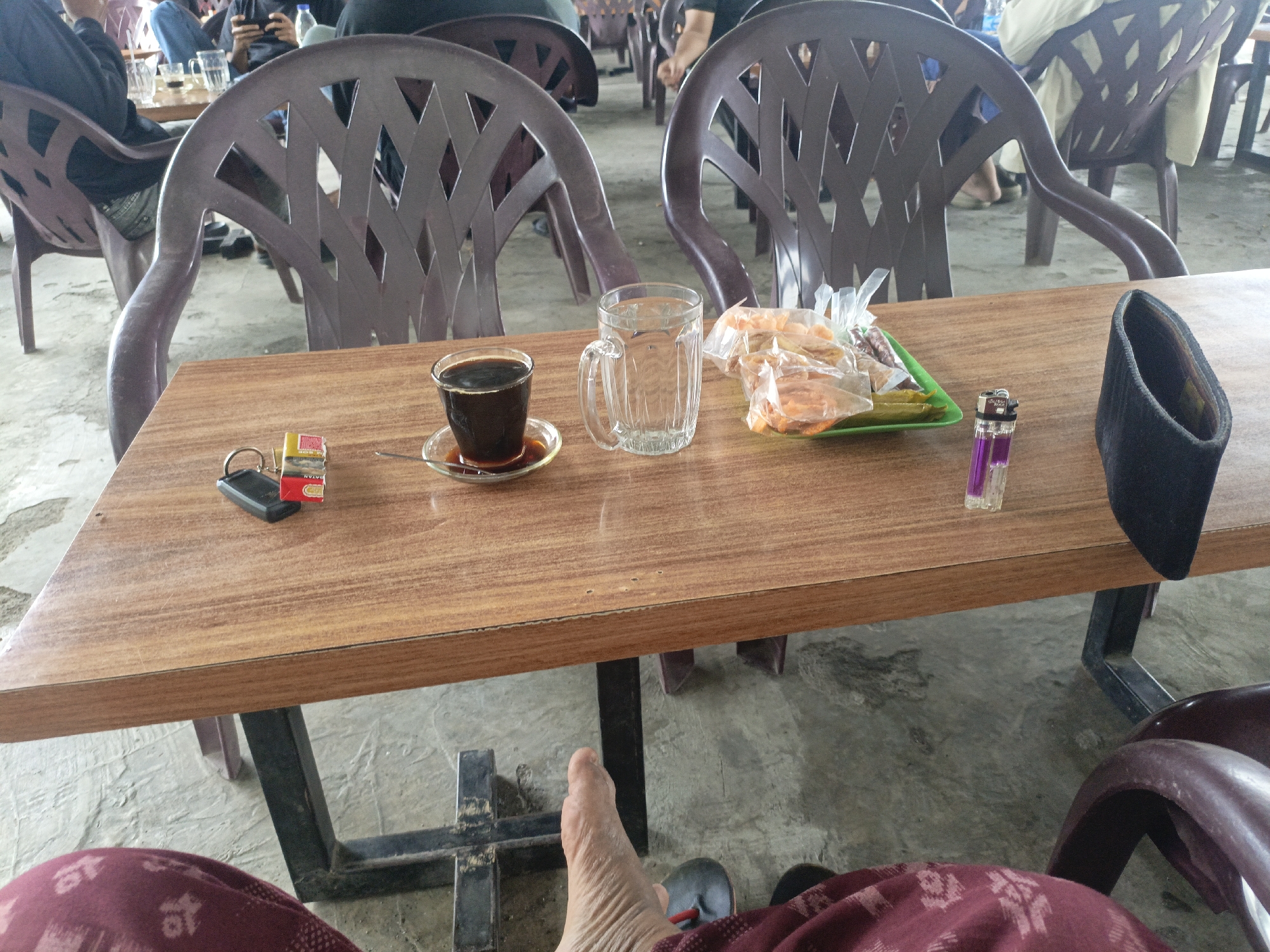 This is a close-up, eye-level shot of a wooden table outdoors, likely at a warung kopi (coffee stall) in Lhokseumawe, Indonesia. The table is positioned between several dark purple plastic chairs, and the ground appears to be a concrete surface with some staining.
On the table, from left to right, are: a small black object with a red and yellow cigarette pack, a glass of dark coffee with a saucer, an empty, fluted glass, a green plastic tray holding several plastic bags of fried snacks, a purple disposable lighter, and a black cloth pouch, possibly a cigarette case or wallet. In the background, blurred figures are seated in chairs, suggesting a public gathering. In the foreground, a bare foot with wrinkled skin is visible, wearing a dark slipper. The person's leg is covered by a maroon fabric with a patterned design. The scene suggests a casual setting, likely a place where people gather for drinks and snacks.