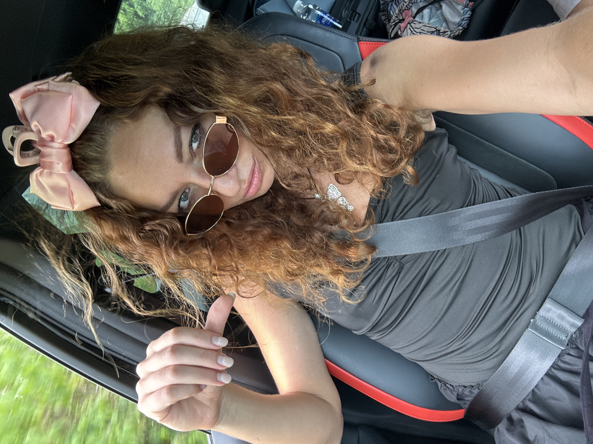 A woman with curly auburn hair, wearing sunglasses and a pink bow in her hair, sits in the driver's seat of a car. She is wearing a black dress and a seatbelt. Her left hand is raised, pointing her index finger towards the camera. The car appears to be in motion, with greenery visible through the window. The interior of the car is dark, with a red accent on the seat. A bottle of water is visible in a cup holder. The scene appears to be a selfie taken by the woman while driving.