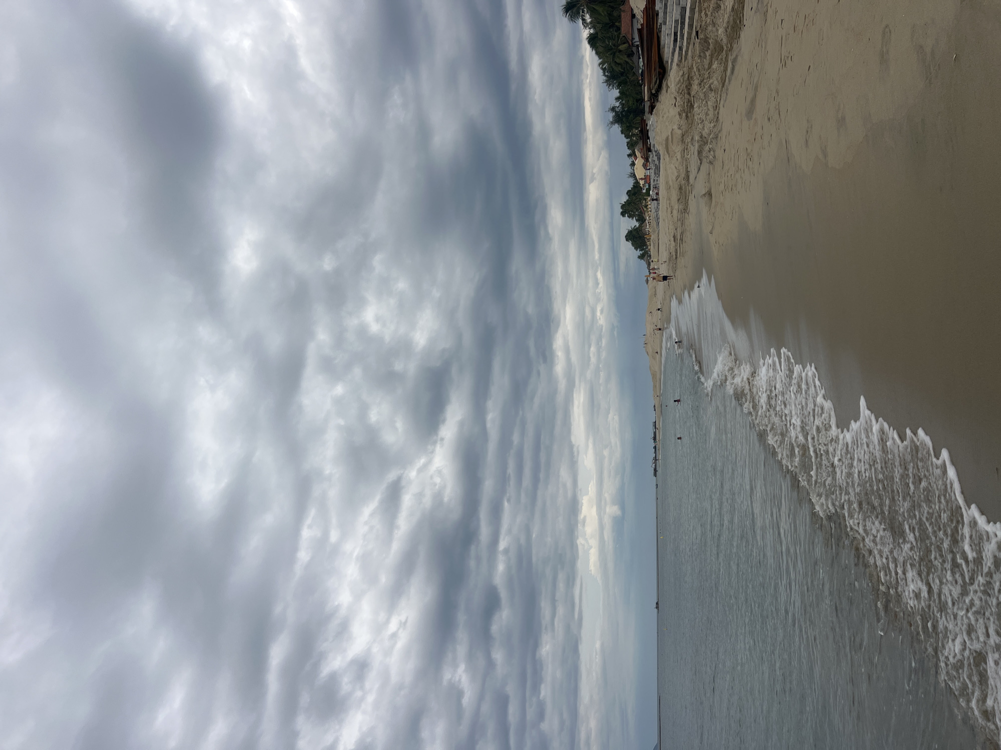 The image depicts a beach scene on a cloudy day, likely in Hoi An, Vietnam, given the presence of palm trees and tropical vegetation along the coastline. The sky is filled with heavy, grey clouds, suggesting an overcast or potentially rainy atmosphere.
The foreground is dominated by a sandy beach. Gentle waves with white foam are lapping at the shore, creating a dynamic border between the water and the sand. The sand appears light brown and slightly wet near the water's edge, becoming drier and lighter further inland.
Along the beach, there are scattered individuals, some walking along the shoreline, others appearing to be swimming or wading in the water. A few small structures, possibly beach umbrellas or huts, are visible further up the beach, as are some boats docked or resting on the sand. The vegetation line behind the beach consists of dense palm trees and other tropical plants, characteristic of a coastal Southeast Asian landscape. In the distance, the sea stretches out to the horizon, meeting the cloudy sky. The overall atmosphere is calm and somewhat subdued due to the weather conditions.