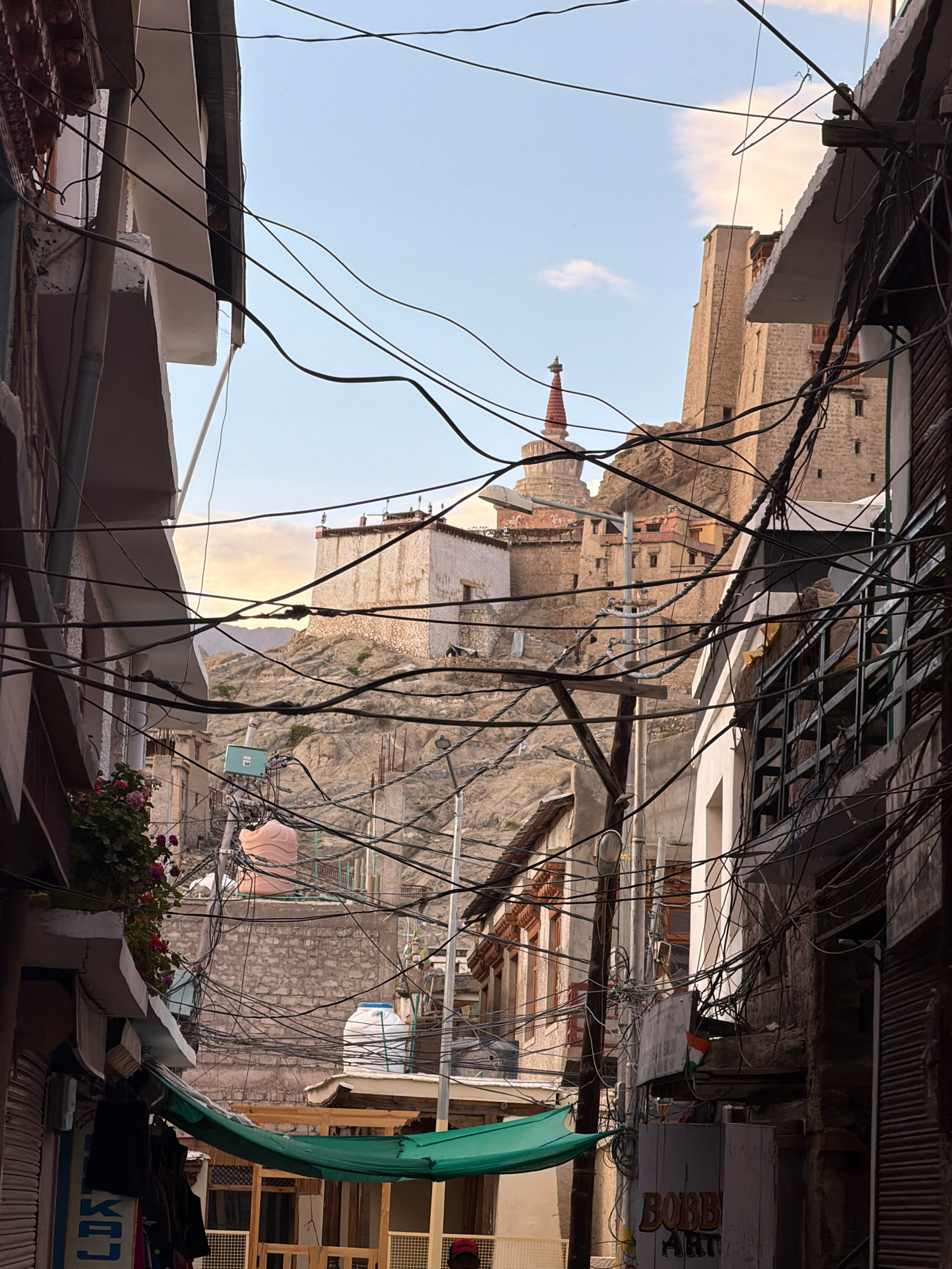 The image depicts a narrow street in Leh, India, likely in the Old Town, with traditional architecture and a prominent monastery or palace complex on a hill in the background. The foreground is dominated by a dense network of overhead electrical wires, a common sight in many Indian cities. Buildings line both sides of the street, some with balconies and shops at ground level. In the background, the Leh Palace or a similar fortress-like structure, characterized by its multi-storied stone construction and battlements, is visible atop a rocky incline. A stupa with a red spire is situated near the palace complex. The sky above is a pale blue with some wispy clouds, suggesting it is either early morning or late afternoon. A green tarp is stretched across part of the street, providing shade. The overall scene conveys a sense of a bustling, lived-in urban environment with a historical and spiritual backdrop.