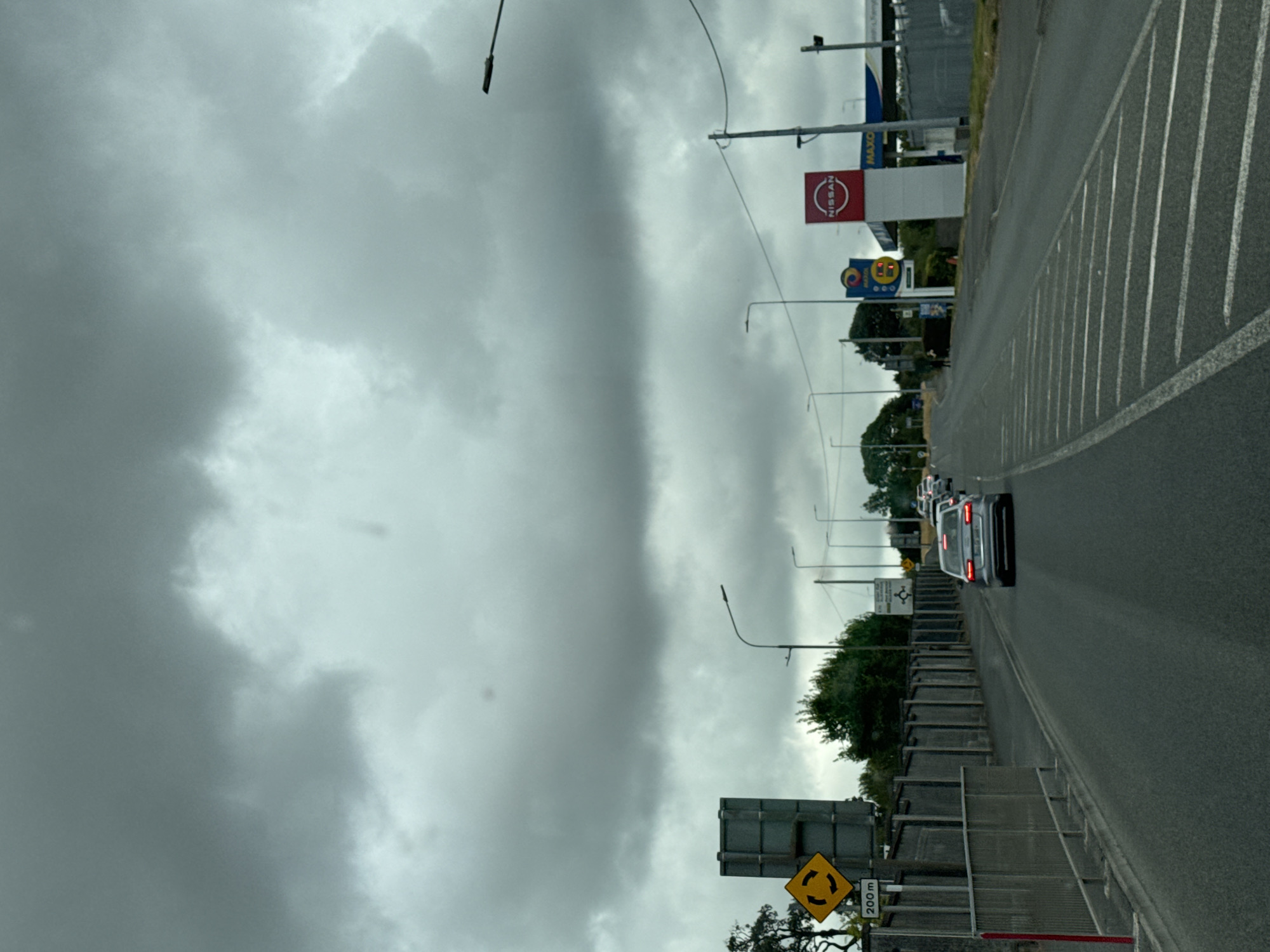 The image captures a view from a vehicle traveling on a road in Port Láirge, Ireland. The sky is overcast with heavy grey clouds. On the left side of the road, a large grey directional sign with a yellow roundabout symbol and "200m" is visible, along with a metal fence and some trees. Several cars are on the road, with the closest one being a silver sedan. On the right side of the road, there are streetlights, a tall white sign with a red Nissan logo, and a blue and yellow Maxol gas station sign. The road has white lane markings, including diagonal lines indicating a merging lane or hazard. The overall impression is of a slightly gloomy day in a suburban or commercial area.