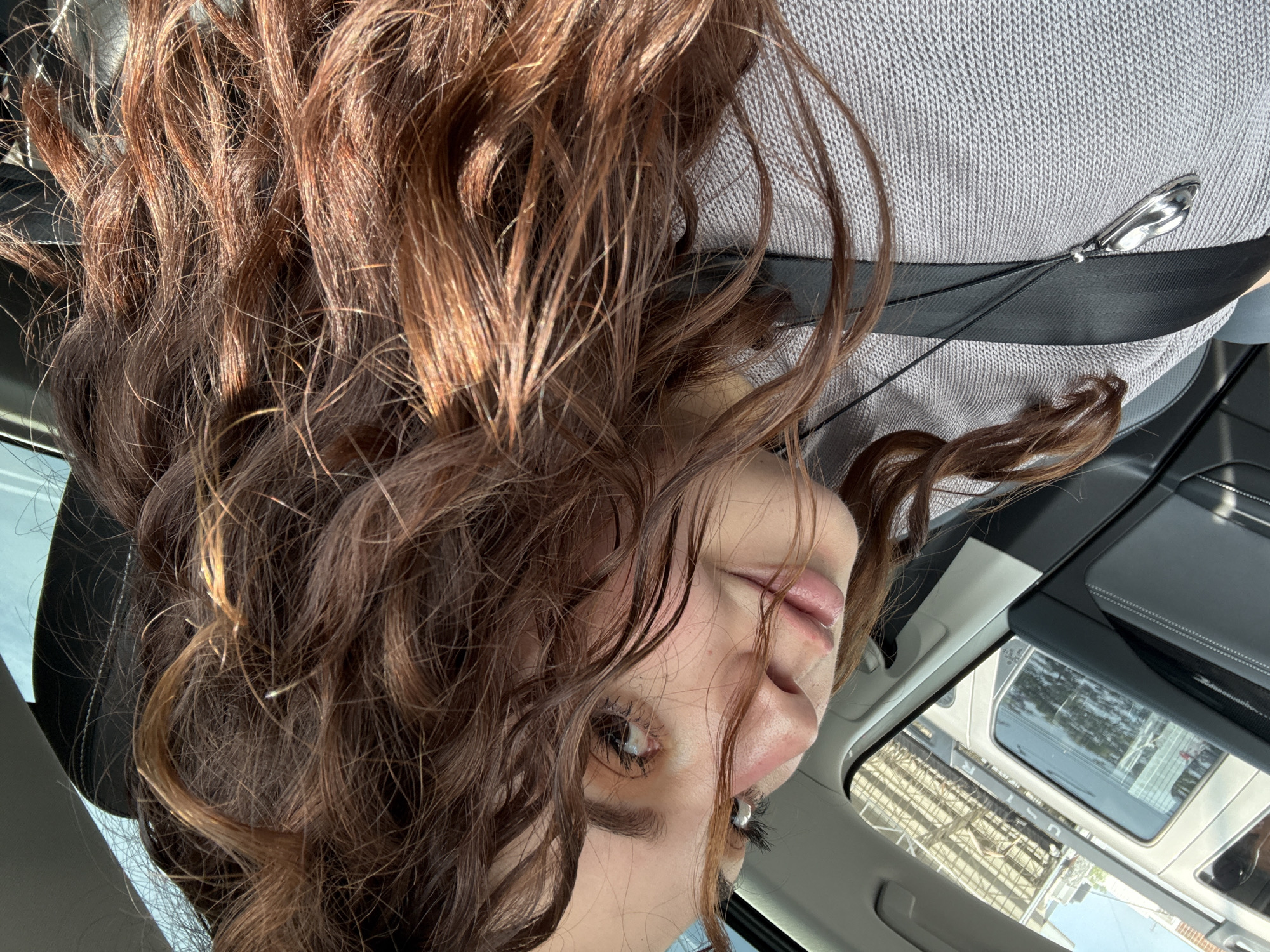A woman with curly brown hair is inside a car, wearing a gray sweater and a black seatbelt with a silver ornament attached. The car has a sunroof, and through it, a view of a city street with other vehicles and buildings is visible. Based on the architecture and vehicles visible through the sunroof, the location is possibly Mexico City, Mexico. The woman appears to be leaning backward or tilting her head down. She has light skin, dark eyes, and visible eyelashes.