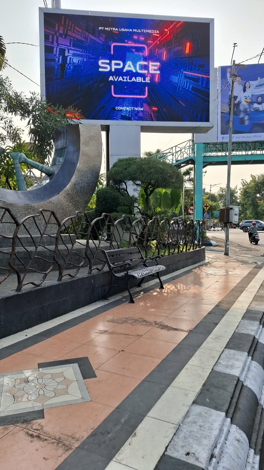 The image depicts an outdoor scene in Kudus, Indonesia, featuring a large digital billboard displaying the words "SPACE AVAILABLE" and "CONTACT NOW" in prominent blue and red lettering against a futuristic background. Below the billboard, a decorative metal fence surrounds a garden area with manicured trees and bushes. A dark metal bench sits in front of the fence, placed on a tiled walkway. The walkway is composed of orange and gray tiles, with a decorative floral pattern tile in the lower left corner. In the background, a street with traffic, including a motorcycle and a car, is visible. An elevated walkway or bridge with a turquoise railing crosses overhead. Another billboard, featuring images of smiling people and a cartoon character, is located to the right of the main digital billboard. The scene is illuminated by natural daylight, casting shadows on the walkway.