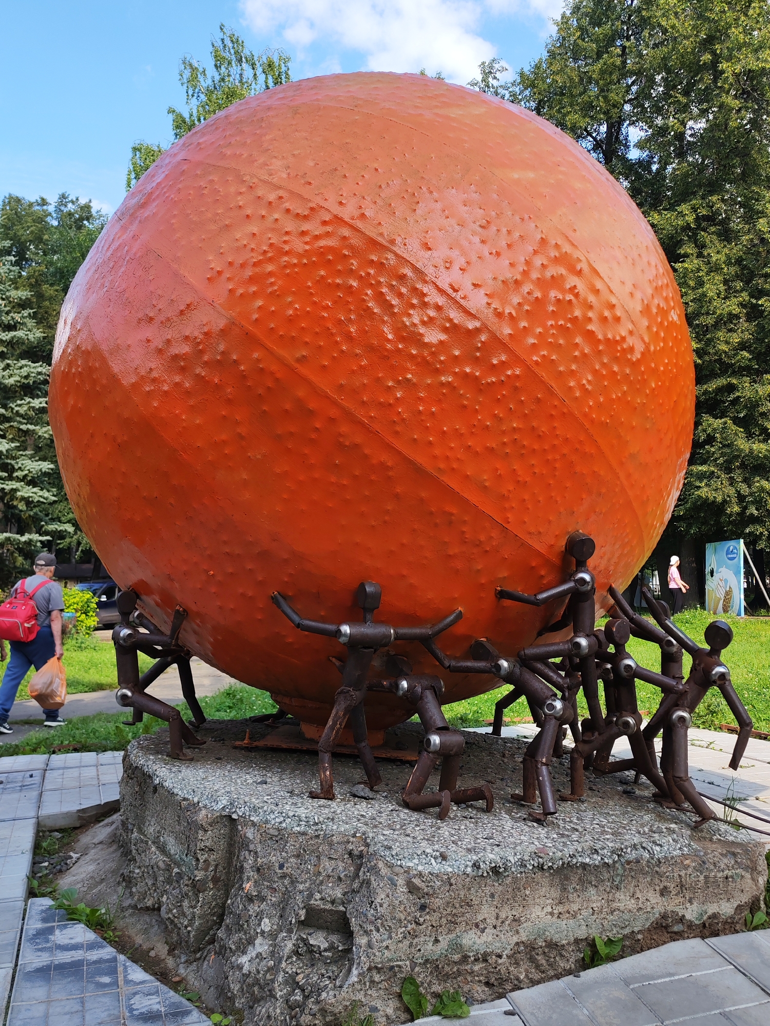 The image shows a large, orange, spherical sculpture with a textured surface, resembling a giant basketball or orange. It is supported by a cluster of small, dark metal figures that appear to be straining to hold it up. The sculpture is situated on a rough, concrete pedestal. The surrounding environment includes a paved walkway with grey tiles, green grass, and lush trees. In the background, a person with a red backpack is walking away from the sculpture, carrying a plastic bag. A distant sign with blue and white imagery is also visible. This sculpture is located in the city of Izhevsk, Russia.