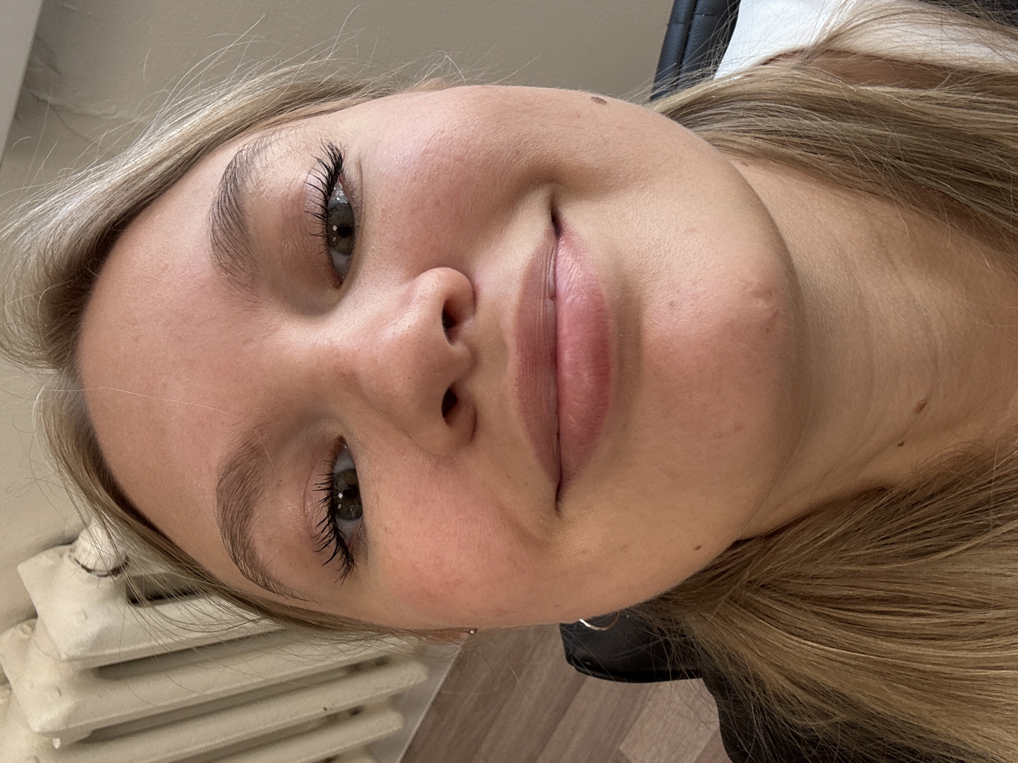 A close-up, slightly rotated portrait captures a young woman with light blonde hair and fair skin, smiling gently at the camera. Her expressive eyes, framed by prominent dark eyelashes, and natural-looking eyebrows are central to her pleasant expression. She has full, naturally colored lips and a few small moles are visible on her forehead and near her right nostril. Her light hair cascades around her face and shoulders. She appears to be wearing a dark top, with a glimpse of a black collar or fabric on her right. The indoor setting includes part of a white, ribbed radiator on the left side of the frame and a wooden floor or surface on the right. The lighting is soft and even, suggesting an indoor environment. There is no discernible activity beyond posing for the photo. No text is visible in the image. The photo was taken in Krasnoyarsk, Russia.