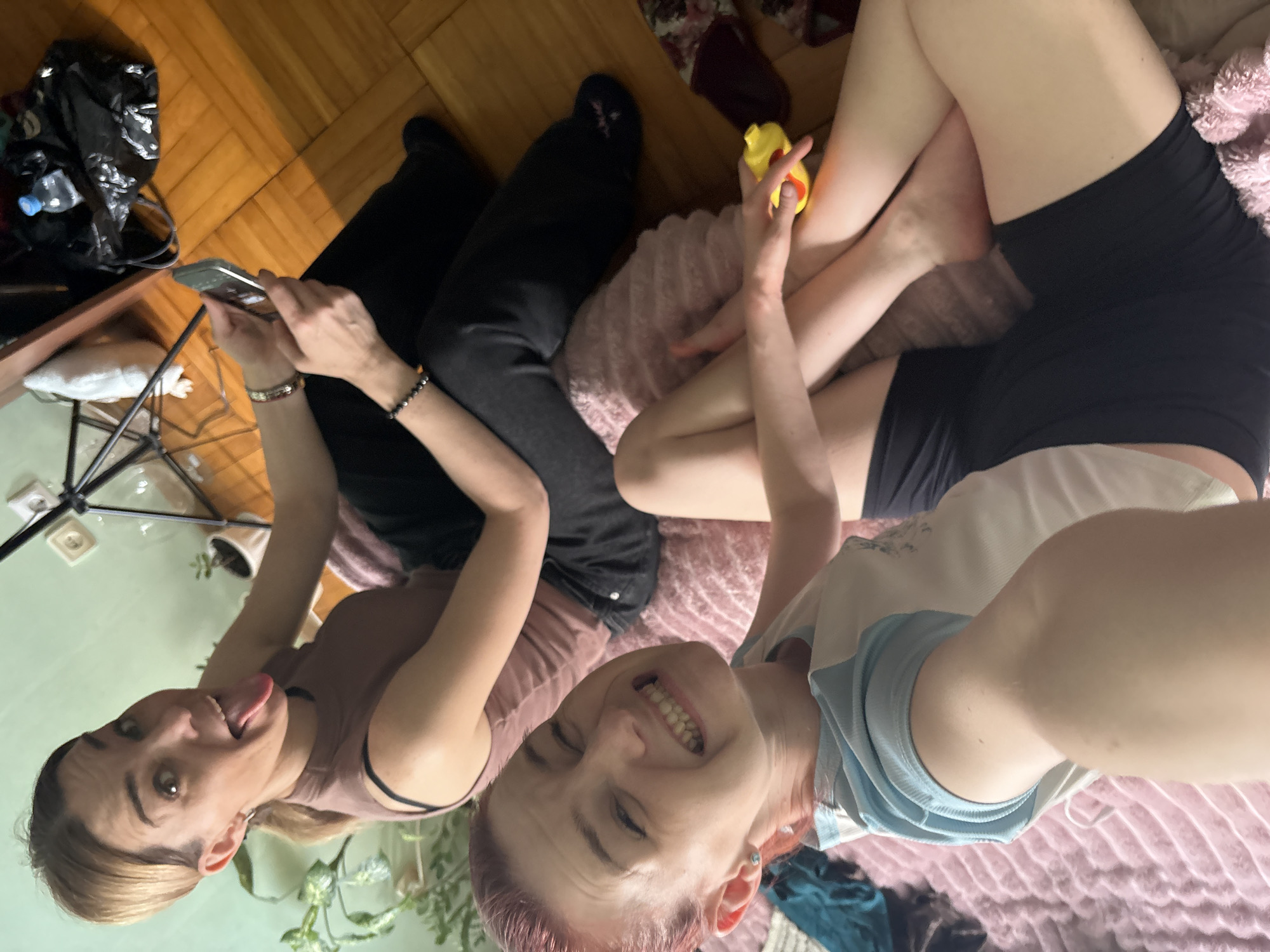 Two women are indoors, likely in an apartment in Vsevolozhsk, Russia, as indicated by the provided context. One woman, with reddish hair pulled back, is in the foreground, holding her phone and making a playful grimace. She is wearing a blue and white t-shirt and black shorts, and is lying on a pink, textured surface. The second woman, with blonde hair in a ponytail, is in the background, looking at her phone. She is wearing a brown tank top and black pants. The environment appears to be a living space with wooden flooring and a light green wall. Objects present include houseplants, a camera tripod, a black trash bag with a water bottle inside, and a yellow toy. The lighting suggests an indoor setting, possibly during the day.