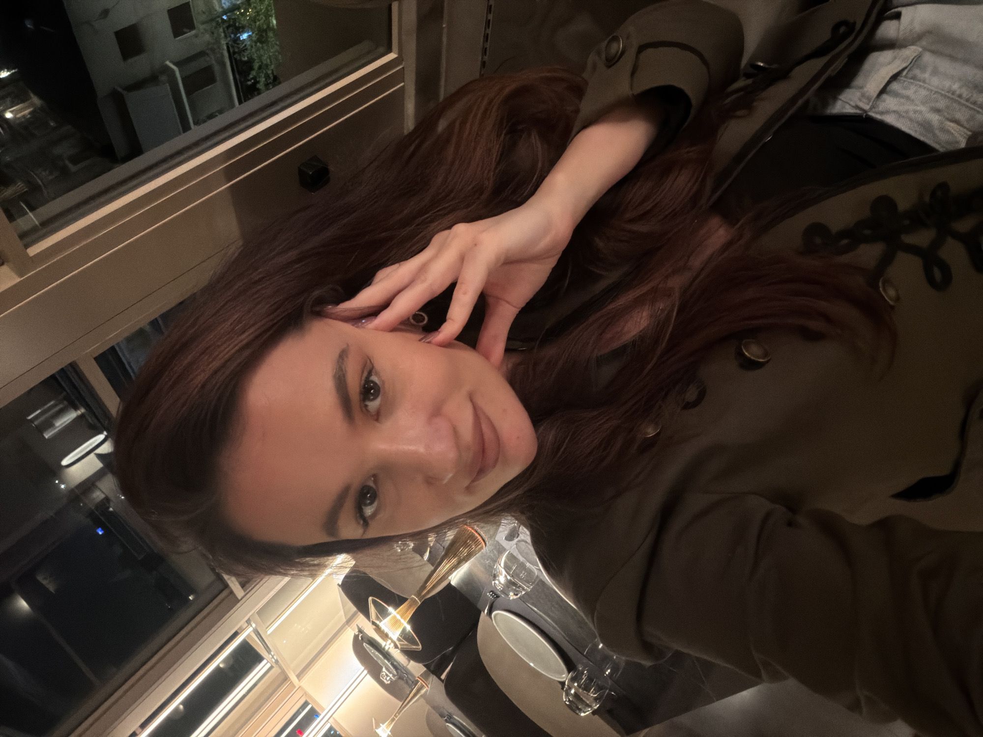 A selfie-style image, rotated 90 degrees clockwise, depicts a young woman posing indoors at night. The woman, with long, dark brown hair, looks directly at the camera with a soft smile. Her right hand is raised, with fingers gently touching her hair near her temple. She is dressed in a dark olive green jacket with distinctive black ornate embroidery and metal buttons, worn over a light blue denim garment.
In front of her, on a dark table surface, there is an elegant tall gold-colored cocktail glass (possibly containing a drink), an empty clear drinking glass, and a white plate. The background reveals an indoor setting, likely a restaurant or bar, characterized by modern decor. Through a large window, a dark urban night scene is visible, featuring illuminated buildings, distant lights, and some greenery. Reflections of indoor lights are also visible on the window glass.
The image conveys a relaxed and pleasant atmosphere, typical of an evening outing. The time of day is clearly night. While the context suggests Athens, Greece, no explicit visual cues within the image confirm this location. There is no visible text.