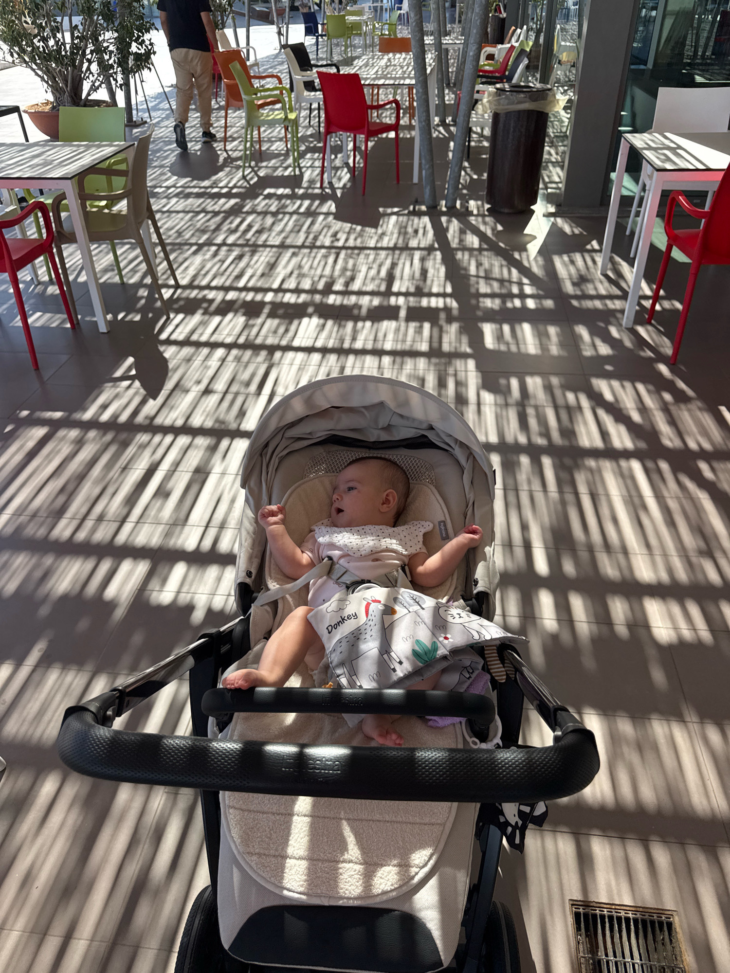 The image captures a baby in a stroller, seated on a tiled patio with strong, linear shadows cast by an overhead structure. The baby, wearing a light-colored garment and a bib with animal prints, is facing left and appears to be looking up. The stroller is beige and black, with a black handlebar in the foreground. In the background, several tables and chairs, predominantly in red, green, and white, are visible, suggesting an outdoor seating area of a cafe or restaurant. A dark cylindrical trash bin stands to the right of a white table. Further in the background, trees and a person wearing khaki pants and a dark t-shirt are discernible. The overall lighting suggests it is daytime with bright sunlight. The location is identified as Isola delle Femmine, Italy.