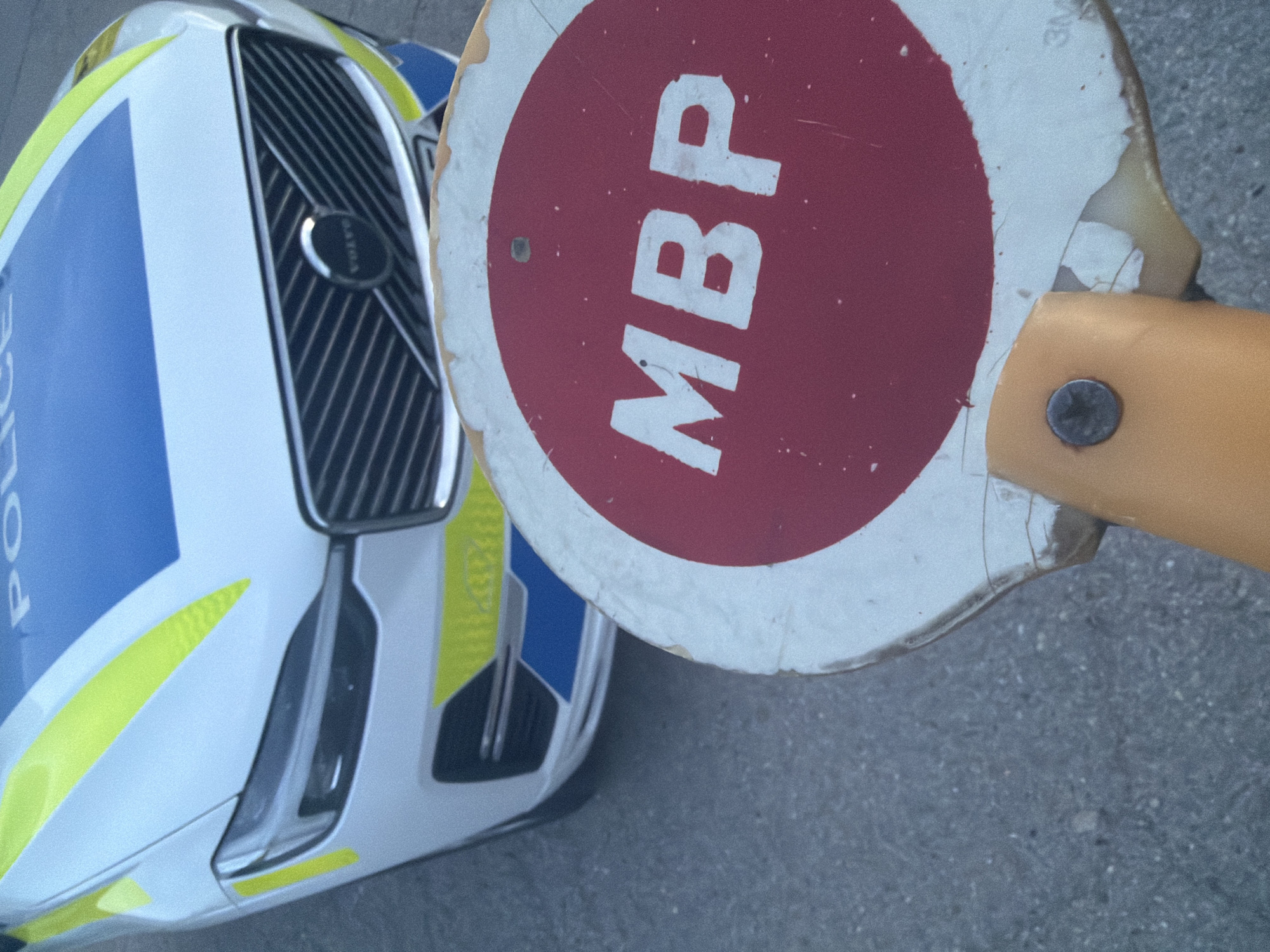 The image shows a close-up, slightly overhead view of the front section of a police car positioned next to a circular handheld sign.
The police car is a Volvo, with its emblem visible on the front grille. Its livery is white, accented with prominent blue sections and fluorescent yellow (or lime green) stripes and patterns. The word "POLICE" is partially visible in white text on the blue section of the car's front.
To the right of the police car is a circular sign featuring a red center and a white outer ring. Bold white capital letters "MBP" are displayed across the red area of the sign. The sign appears worn, with visible chips and scuff marks on its edges and surface. It is attached to a yellow handle or pole, with a metal screw securing it.
The scene is set outdoors on what appears to be a grey, paved surface, likely asphalt. The lighting indicates it is daytime. The combination of the police vehicle and the "MBP" sign (often associated with police or interior ministry functions, particularly in a Bulgarian context) suggests an activity related to law enforcement, such as a checkpoint or traffic control. The location context is Smyadovo, Bulgaria.