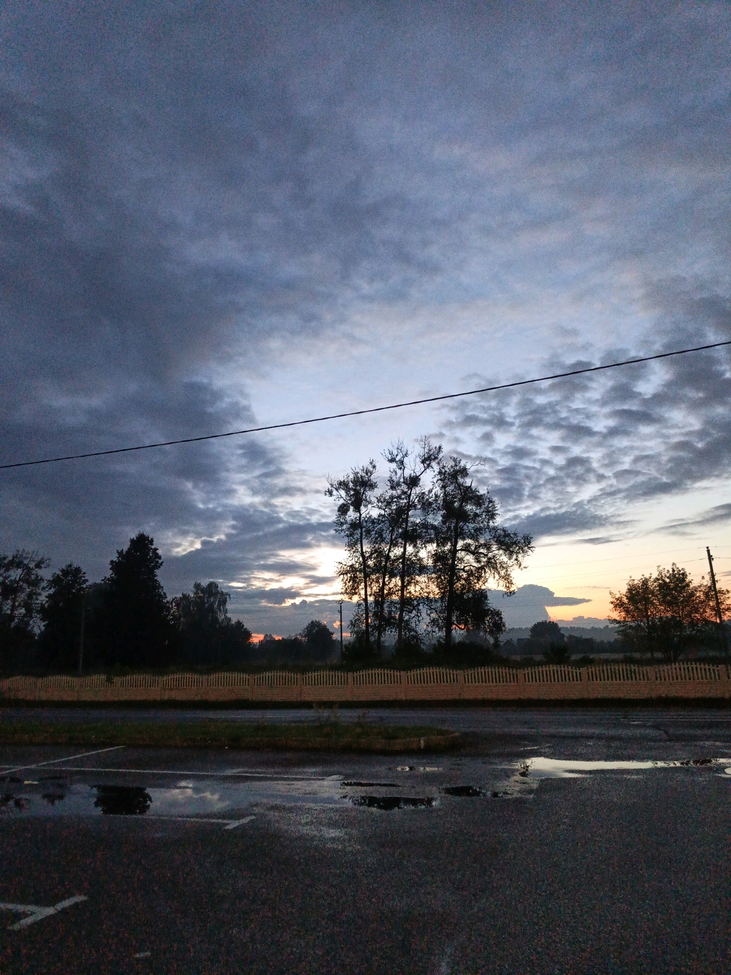 The image depicts a twilight scene in the city of Парэчча, Belarus. The sky is filled with dramatic, dark clouds with streaks of lighter gray and hints of orange and yellow near the horizon, suggesting the last rays of the sun. Silhouetted trees, some bare and others with foliage, line the background, creating a stark contrast against the sky. In the foreground, a dark asphalt surface is visible, with several puddles reflecting the dimly lit environment. A light-colored, wooden picket fence runs horizontally across the middle ground, separating the road or parking area from the natural landscape. A single power line stretches across the upper portion of the frame. The overall atmosphere is serene and somewhat somber, characteristic of dusk or dawn. There are no people or animals visible in the scene.