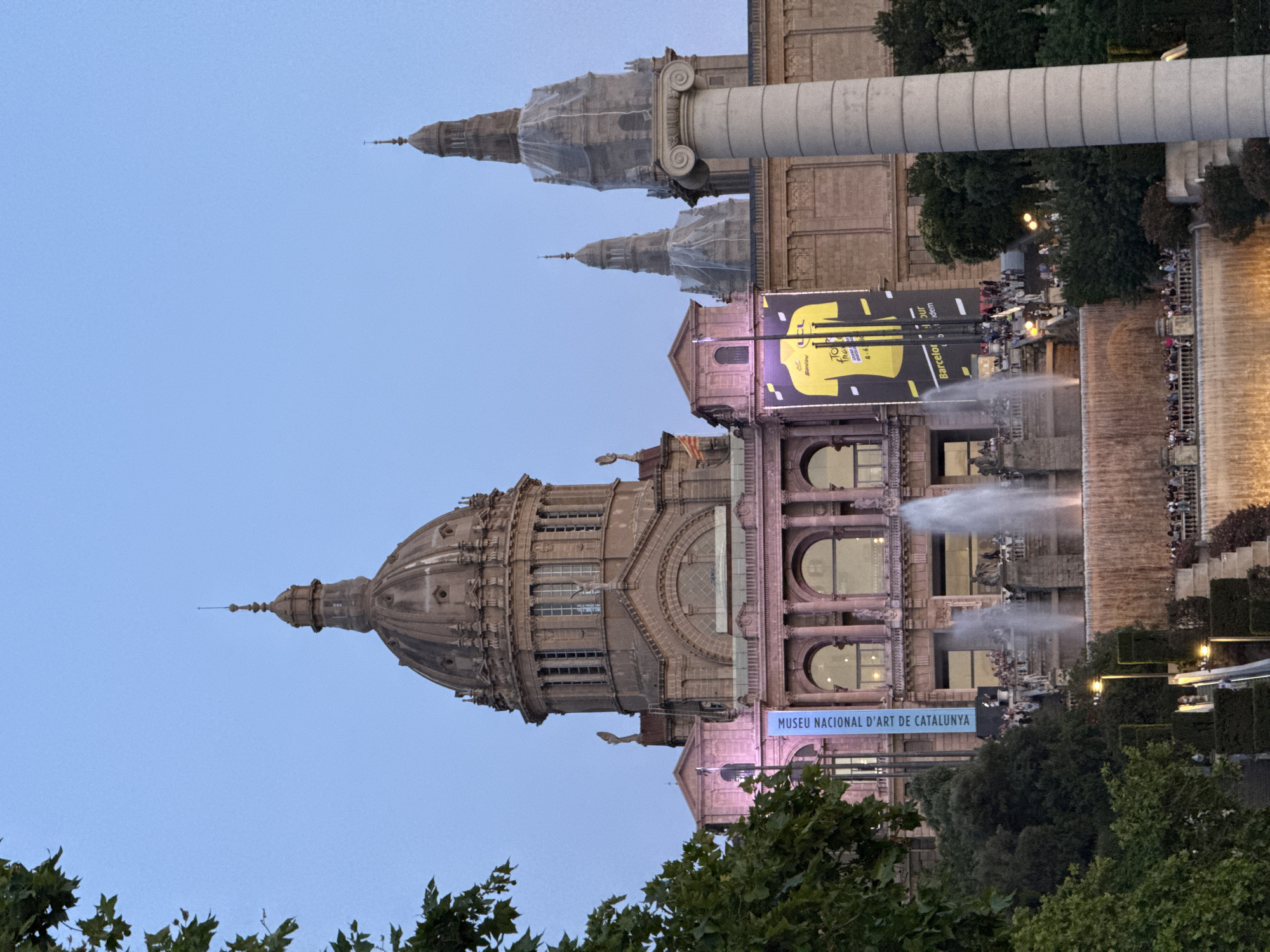 The image shows the Palau Nacional in Barcelona, Spain, an imposing building with a large central dome and two smaller domes. The building is illuminated by pink and yellow lights, and a large banner hangs from its facade. In front of the building, there are tiered fountains with water cascading down, and a crowd of people are gathered on the terraces and steps. The sky is a clear blue, suggesting it is either early morning or late evening. The National Palace is located on Montjuïc hill, overlooking the city of Barcelona.