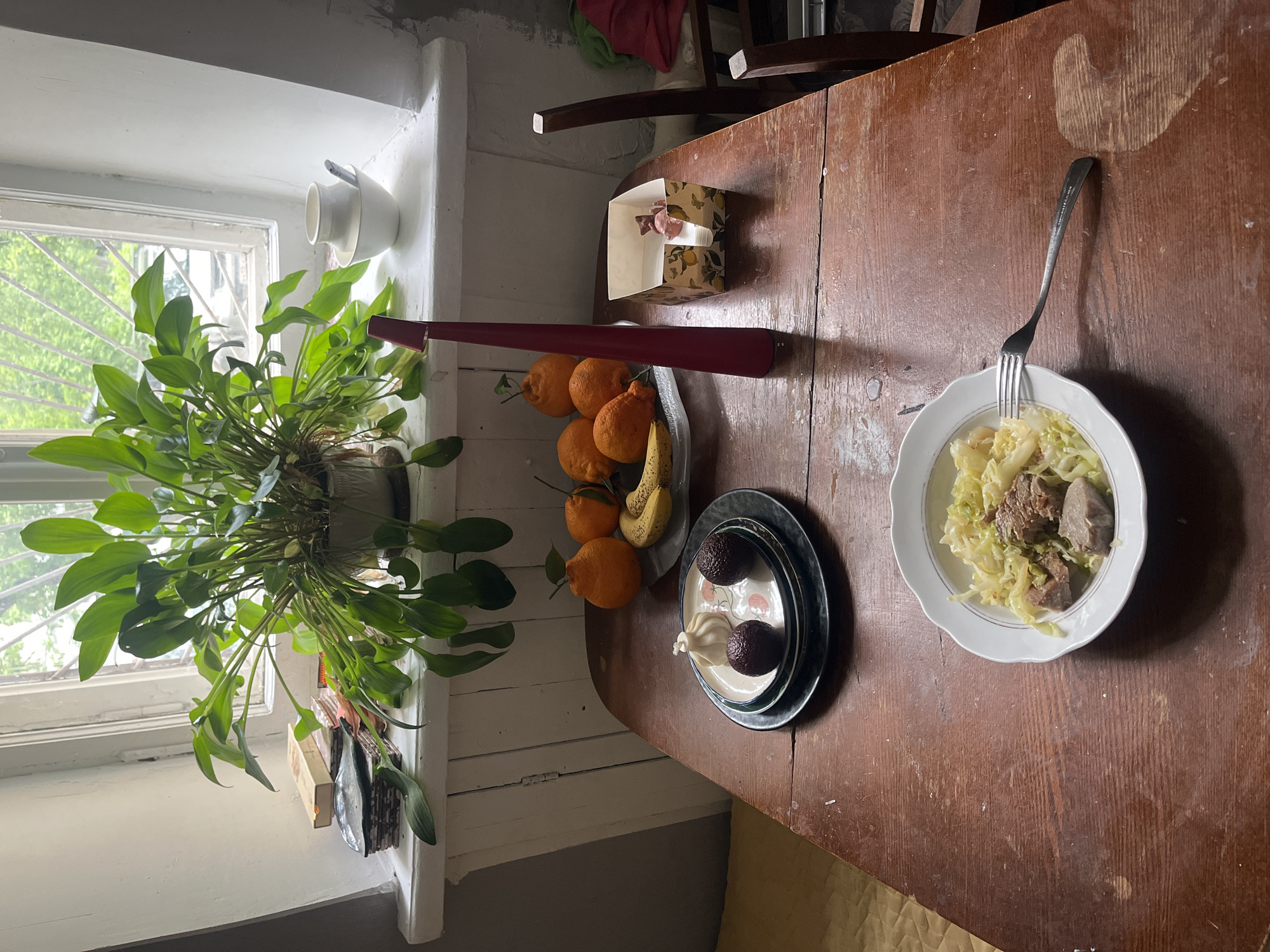 A medium, overhead shot shows a wooden table next to a window with a plant in it. On the table, a plate of cooked cabbage with meat sits on the right side. Adjacent to it, closer to the window, is a plate with two dark, round objects and a small, light-colored object. A bowl of oranges and a banana are situated near a long, red object and a small, patterned box. The scene appears to be indoors. It is not possible to determine if the location is in Перовское сельское поселение, Ukraine based on the visual content.
