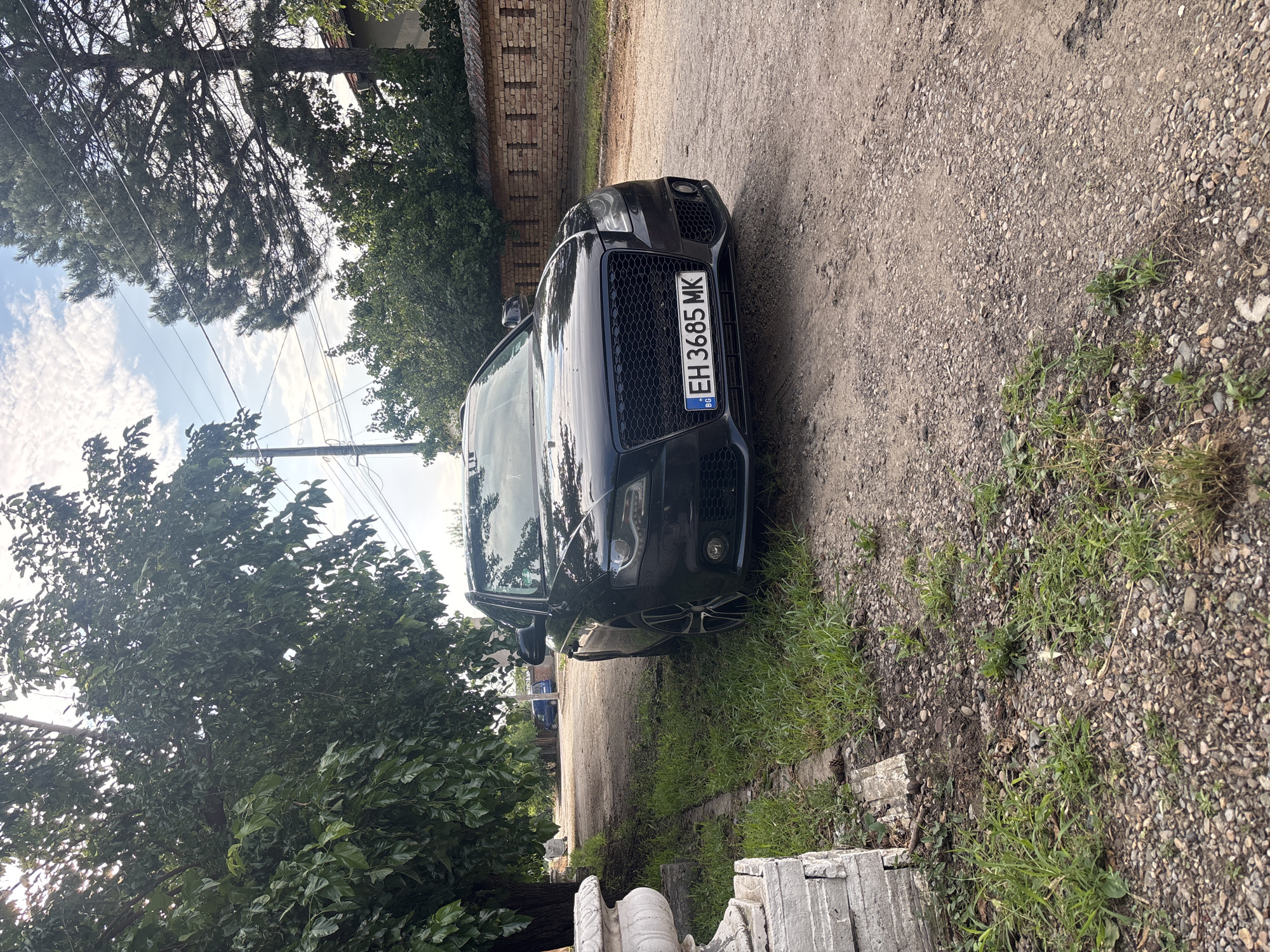 In Brenitsa, Bulgaria, a black Audi A4 sedan is parked on a gravel road lined with grass and trees. The car's license plate reads "EH 3685 MK" with the Bulgarian flag emblem. A white stone planter sits to the left of the car, next to a pile of stacked concrete blocks. In the background, a brick wall, more trees, and a glimpse of a blue vehicle suggest a residential area. The sky is partly cloudy with a bright sun.