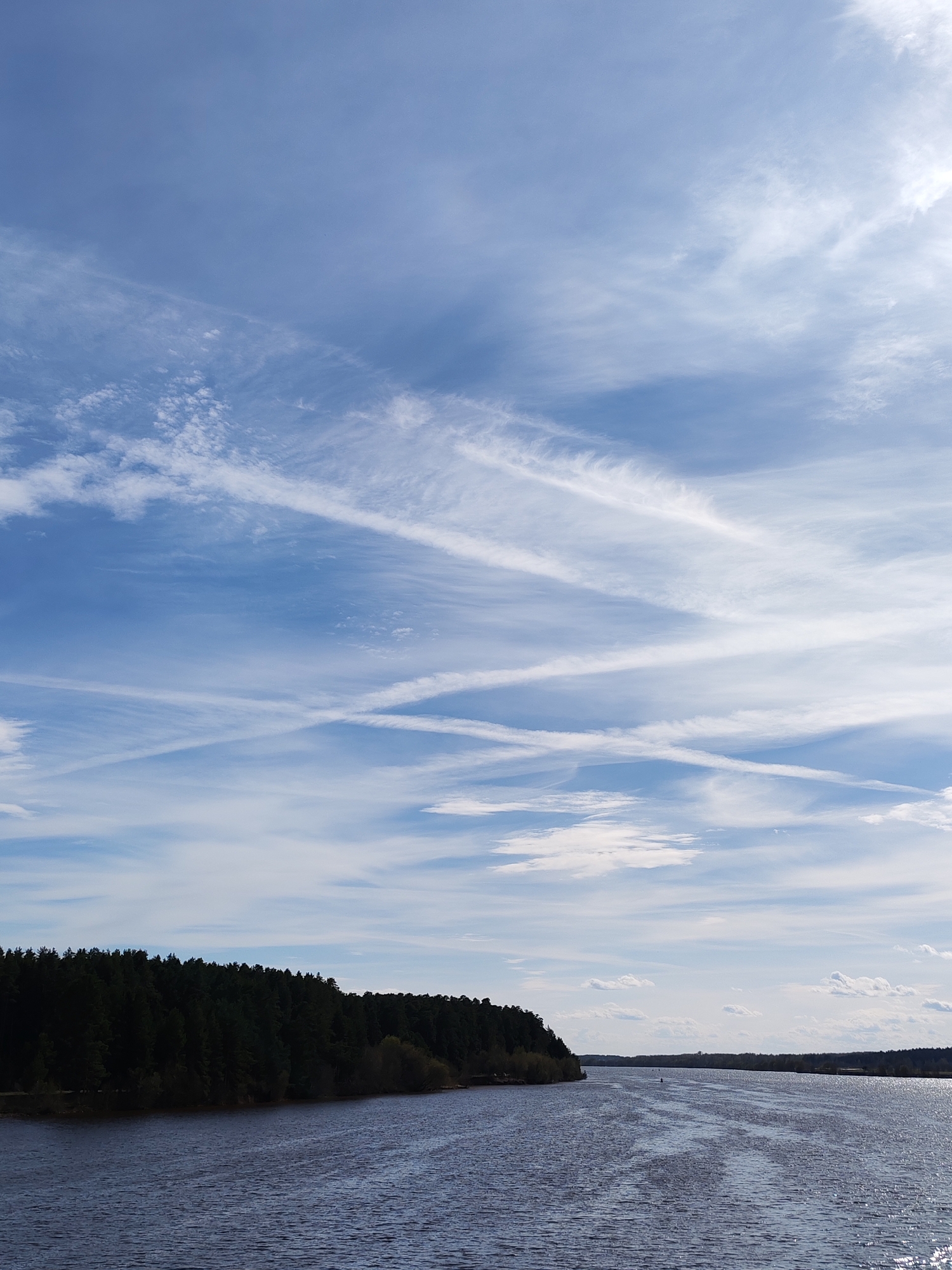 The image depicts a body of water bordered by a densely forested area under a blue sky with numerous contrails. The location is Unknown, Unknown. The water is dark and shows evidence of ripples or waves, possibly from wind or boats. The forest along the shore is comprised of dark green trees, suggesting a coniferous or evergreen species. The sky is mostly blue, interspersed with thin, white contrails. The light suggests it is daytime, though the exact time is indeterminable. There are no visible people.