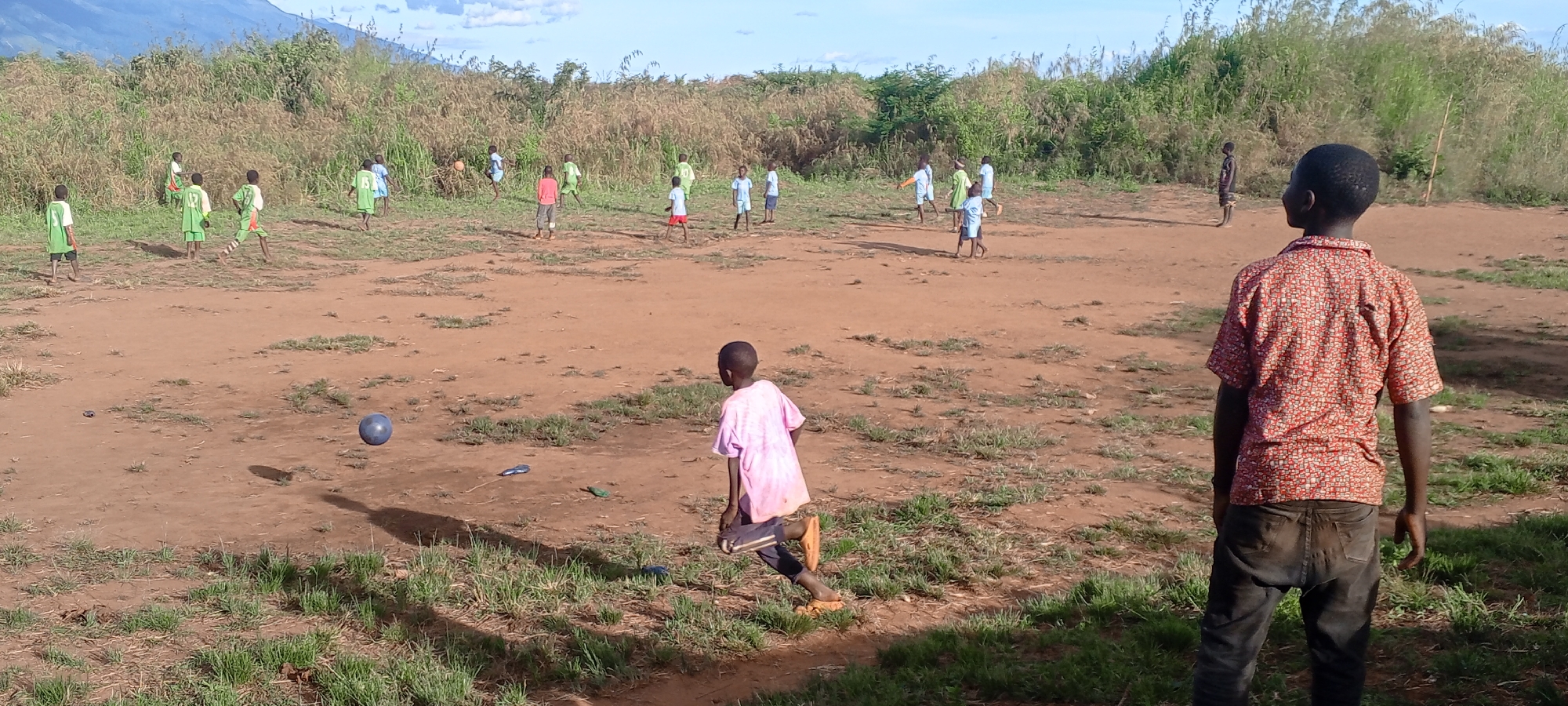An outdoor wide shot captures a lively game of soccer played by numerous children on a reddish-brown dirt field in Ngalula, Democratic Republic of the Congo. The field is uneven, with patches of green grass, and is flanked by tall, dense green brush and grasses. Distant hills or mountains are visible under a partly cloudy blue sky.
Approximately 15 children, mostly boys, are scattered across the field, actively engaged in the game. In the lower left foreground, a dark blue soccer ball is mid-air, casting a shadow, as a child in a pink t-shirt and dark shorts runs towards it, seemingly barefoot or in sandals. To the far right foreground, a boy with his back to the viewer watches the game, wearing a red and white patterned short-sleeved shirt and dark pants.
In the mid-ground, several children wear light green jerseys with numbers on their backs, such as "12" and "15," suggesting team uniforms. Other children wear light blue shirts, a red shirt, or various casual attire. The time of day appears to be sunny daylight, likely late morning or afternoon, given the clear shadows cast by the children and the ball. The scene conveys a sense of energetic, informal play in a natural, rural environment. No other significant text is visible.