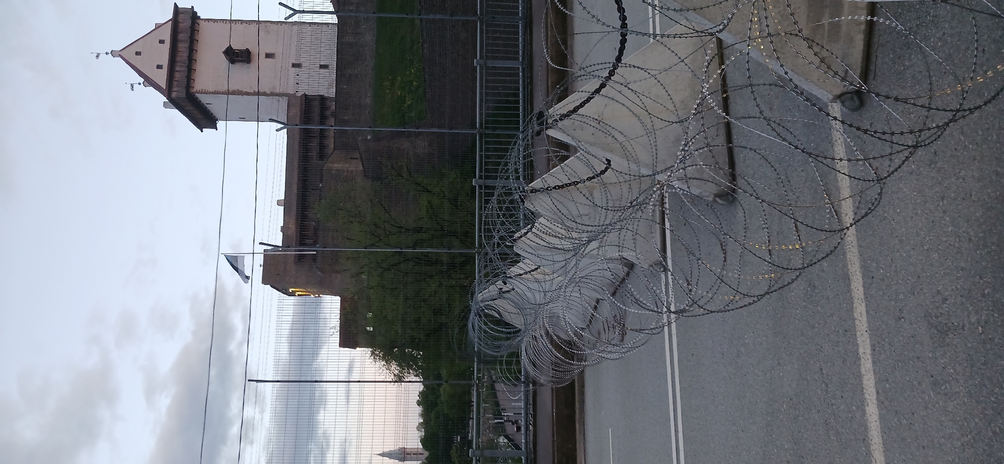 The image depicts a segment of road barricaded with razor wire in Ivangorod, Russia. In the background, the Ivangorod Fortress is visible against a cloudy sky. A metal fence runs parallel to the road, and the Estonian flag is seen. There is no visible human activity.