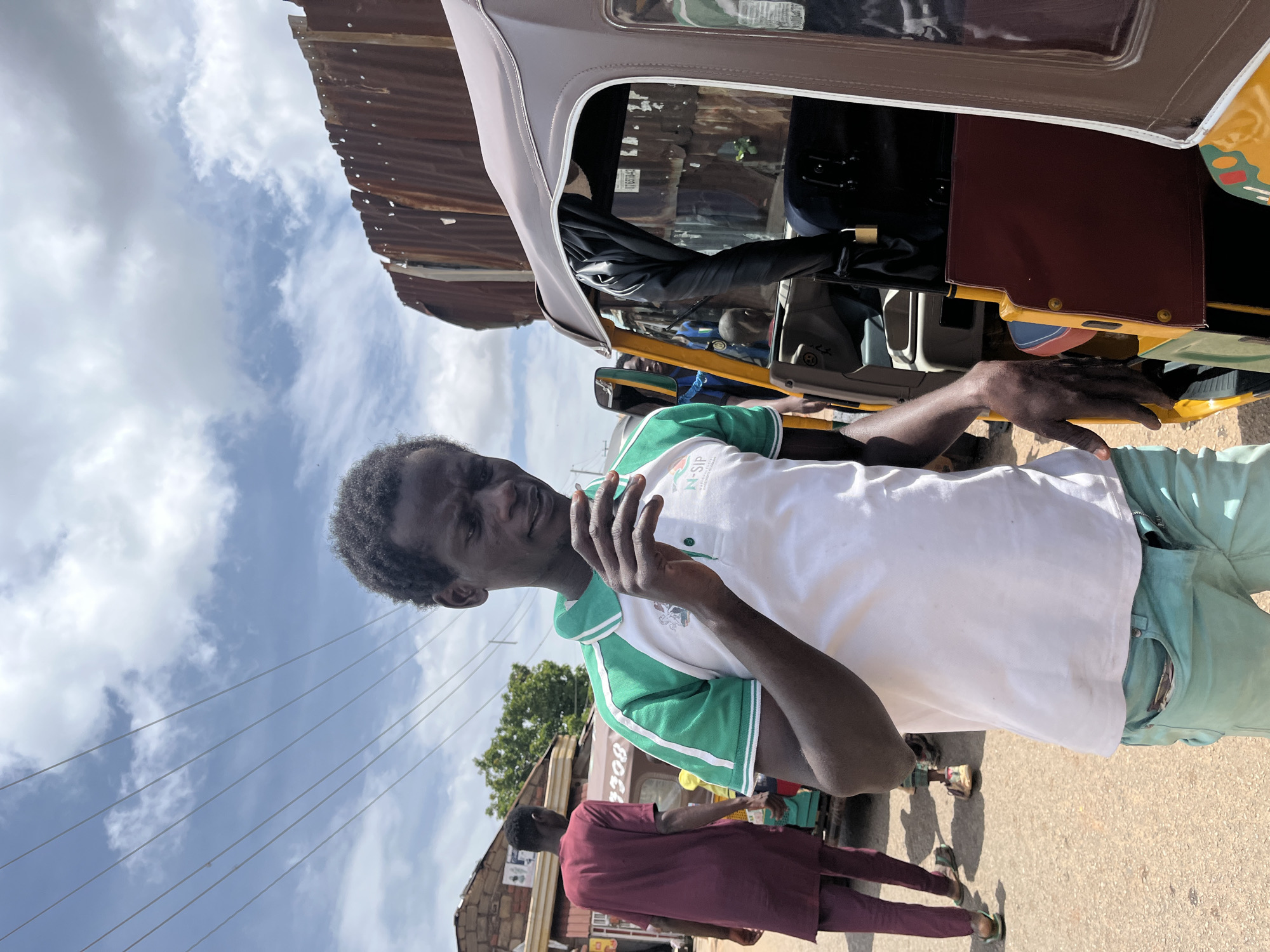 In Gura Top, Nigeria, under a partly cloudy sky, a young Black man stands beside a colorful auto-rickshaw. He wears a white polo shirt with green trim and teal pants. His left hand rests on the auto-rickshaw's door, and he appears to be smoking or holding a small object in his right hand near his mouth. Behind him, another Black man in a maroon outfit walks away, and other auto-rickshaws and buildings are visible in the background. The ground is paved, and electrical wires crisscross the sky.