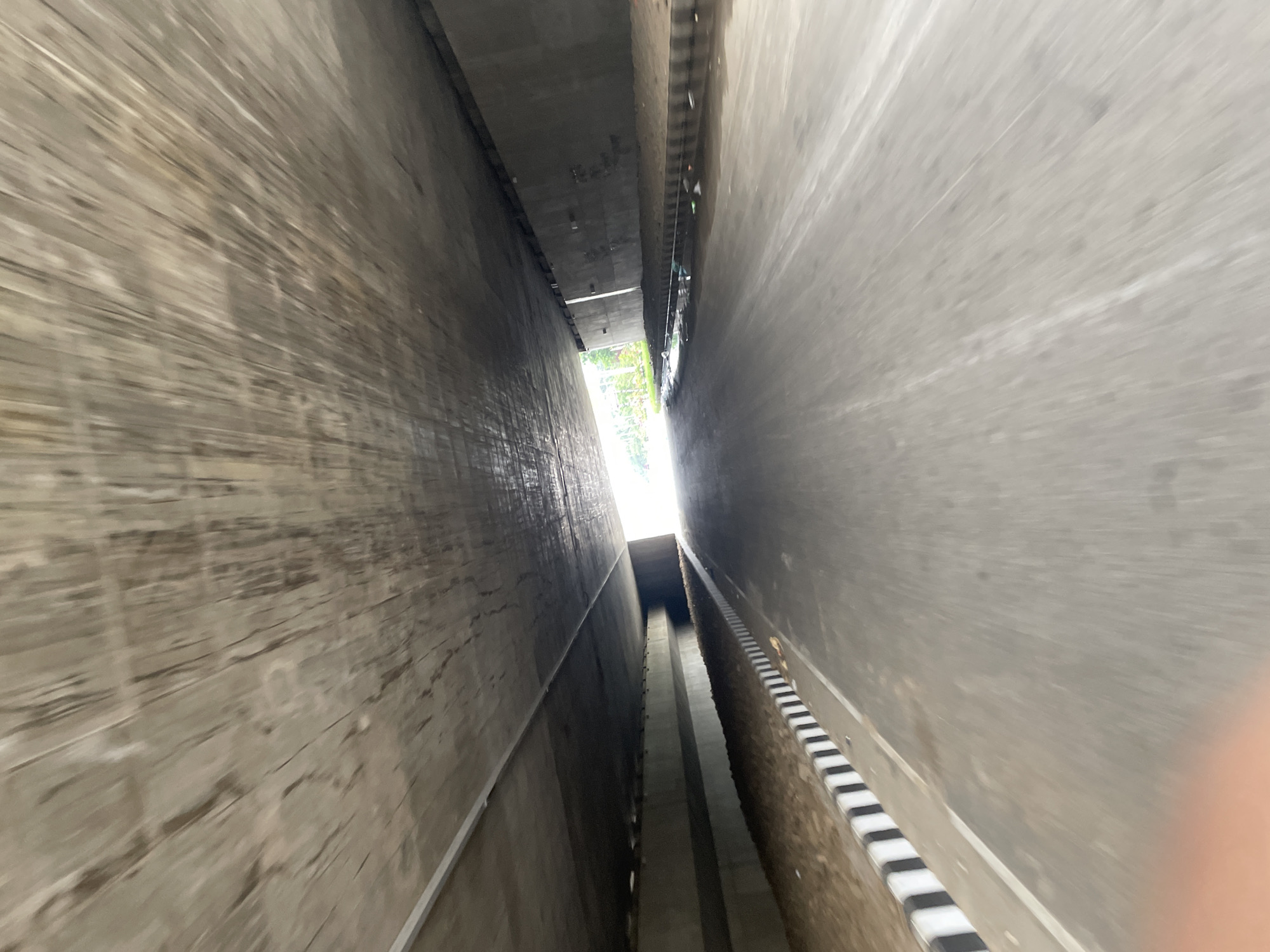 The image depicts a narrow channel with concrete walls in Purbachal, Bangladesh. The channel appears to be a storm water drainage system or a similar type of infrastructure. The walls are tall and close together, creating a deep, confined space. Light is visible at the end of the channel, suggesting an opening or exit point. The walls have varying textures and colors, with some parts appearing aged or stained. A black and white measuring marker is visible on one side of the channel.