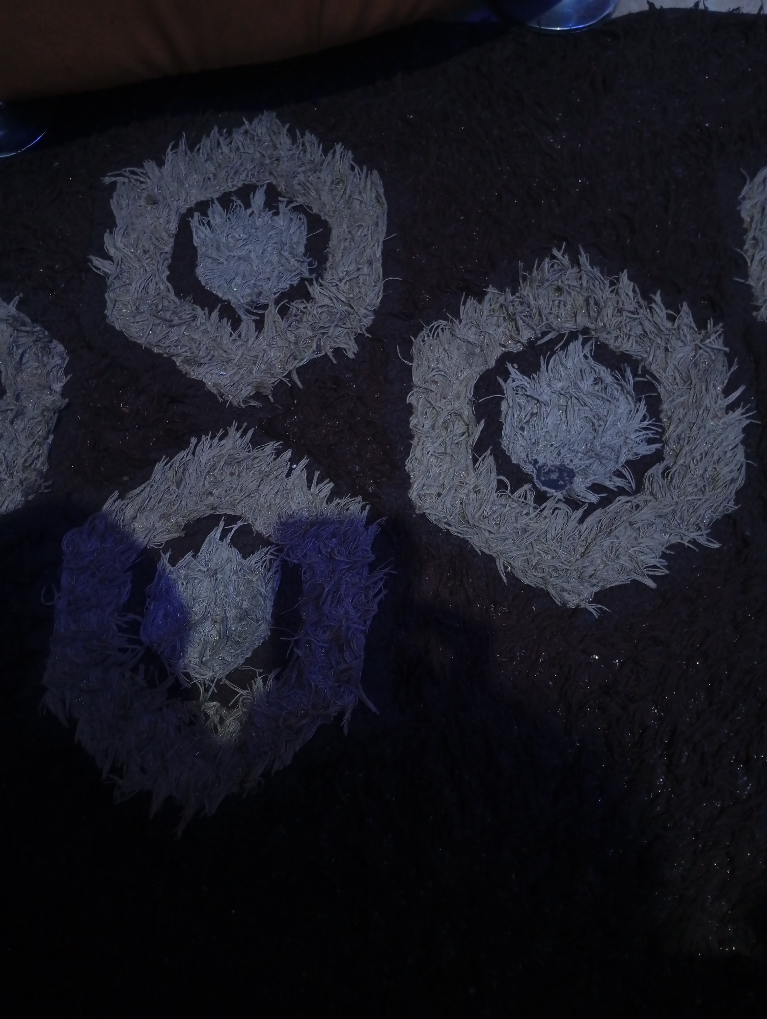 The image displays a close-up view of a dark-colored, shaggy rug or mat, likely situated indoors in Lokoja, Nigeria. The rug features several distinct, lighter-colored patterns against a dark brown or black background.
The patterns are intricately designed, appearing as nested, shaggy shapes that resemble hexagons or circles. Each pattern consists of dense, fibrous material, possibly yarn, forming a raised, high-pile texture. The primary color of these patterns is a light grey or off-white. One of the patterns, located in the bottom-left portion of the image, exhibits a noticeable blue or purple hue, suggesting it might be reflecting ambient light from an artificial source or a nearby object.
The overall lighting in the scene is dim, casting a somewhat moody atmosphere. There are subtle glints of light reflecting off the individual fibers of the rug, particularly on the raised patterns, indicating the presence of some light source, possibly artificial, illuminating the surface. The background of the rug also has a shaggy texture but is less defined due to the darkness. In the upper-left corner, a sliver of what appears to be a piece of furniture, possibly a sofa or chair, is visible, along with a metallic reflection.
No specific activity or event is taking place; the image is a static shot focused on the texture and design of the floor covering. There is no visible text.