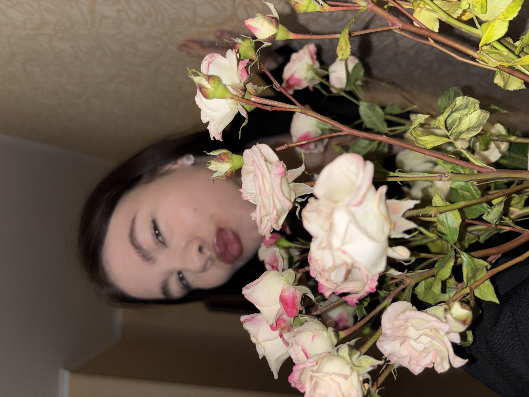 A photograph shows a woman with dark hair and fair skin holding a bouquet of roses. The roses are primarily white with pink edges. Some of the leaves and stems of the roses appear dry. The background appears to be a wall. This image was taken in Perm, Russia.