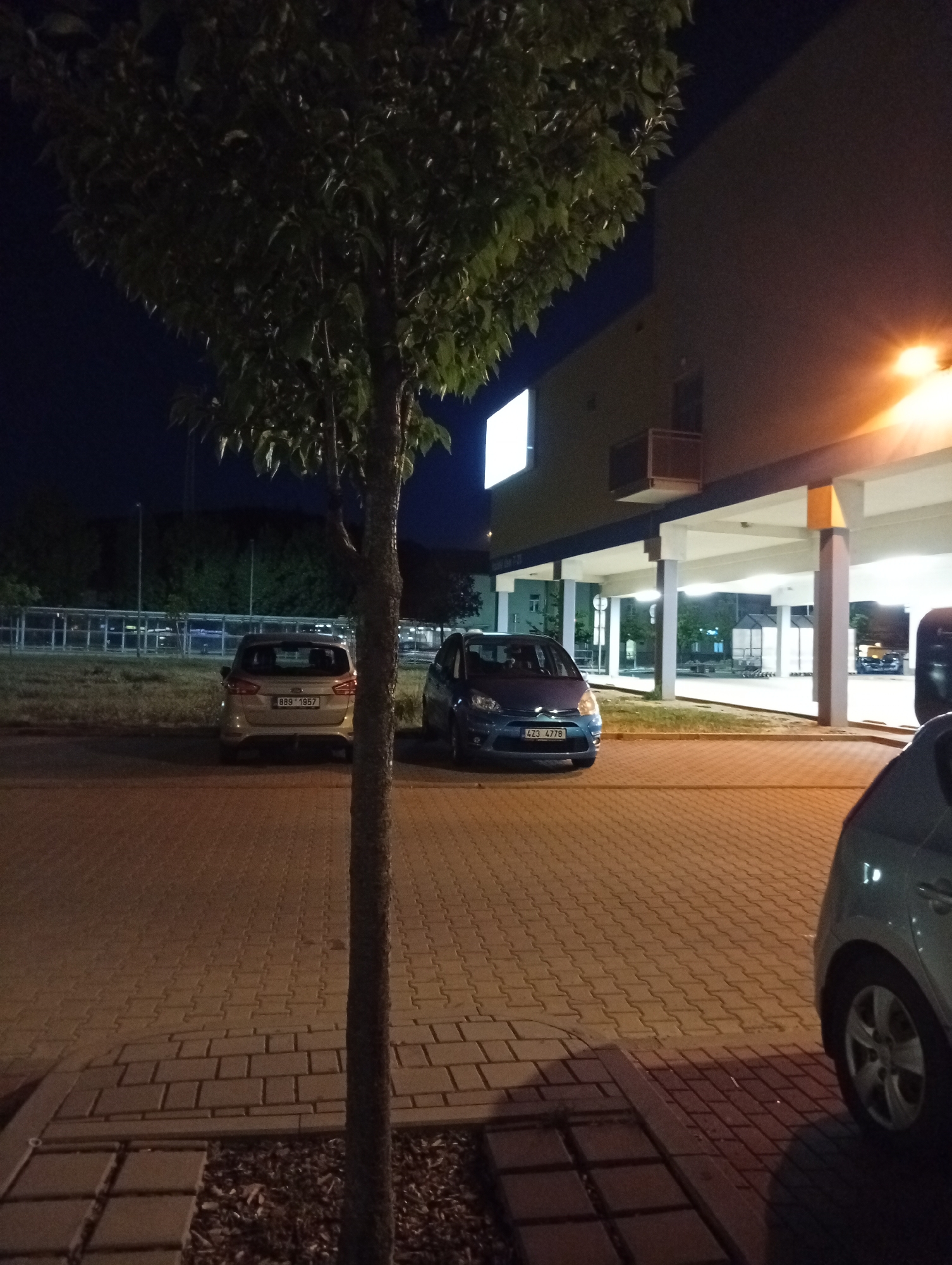 An outdoor, nighttime, medium shot captures a paved parking lot in front of a building in Tišnov, Czechia. A young tree stands in the foreground, its trunk partially obscuring a light-colored car parked to the left. The car's license plate reads "B89 1957". To the right of the light-colored car, a blue hatchback is parked, its license plate displaying "423 4778". The blue car's headlights are on. In the background, a building with covered walkways and pillars is visible, illuminated by artificial lights. The sky above is dark. The ground is covered in light-colored interlocking pavers, with some areas of grass and mulch visible. The overall scene suggests a typical urban parking area after dark.