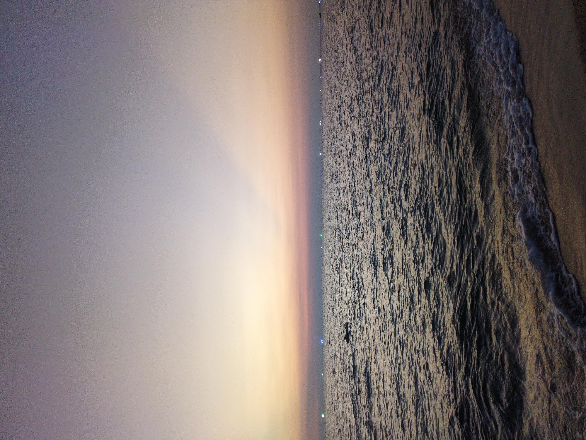 The image depicts a coastal scene in Phú Quốc, Vietnam, taken at twilight or dawn. A smooth gradient sky transitions from a darker hue at the top left to a lighter, golden color near the center horizon. Below this horizon, a body of water extends to the right. Gentle waves are breaking on the sand. There are some bright lights that appear to be from buildings in the distance. A small boat is visible in the center of the sea.
