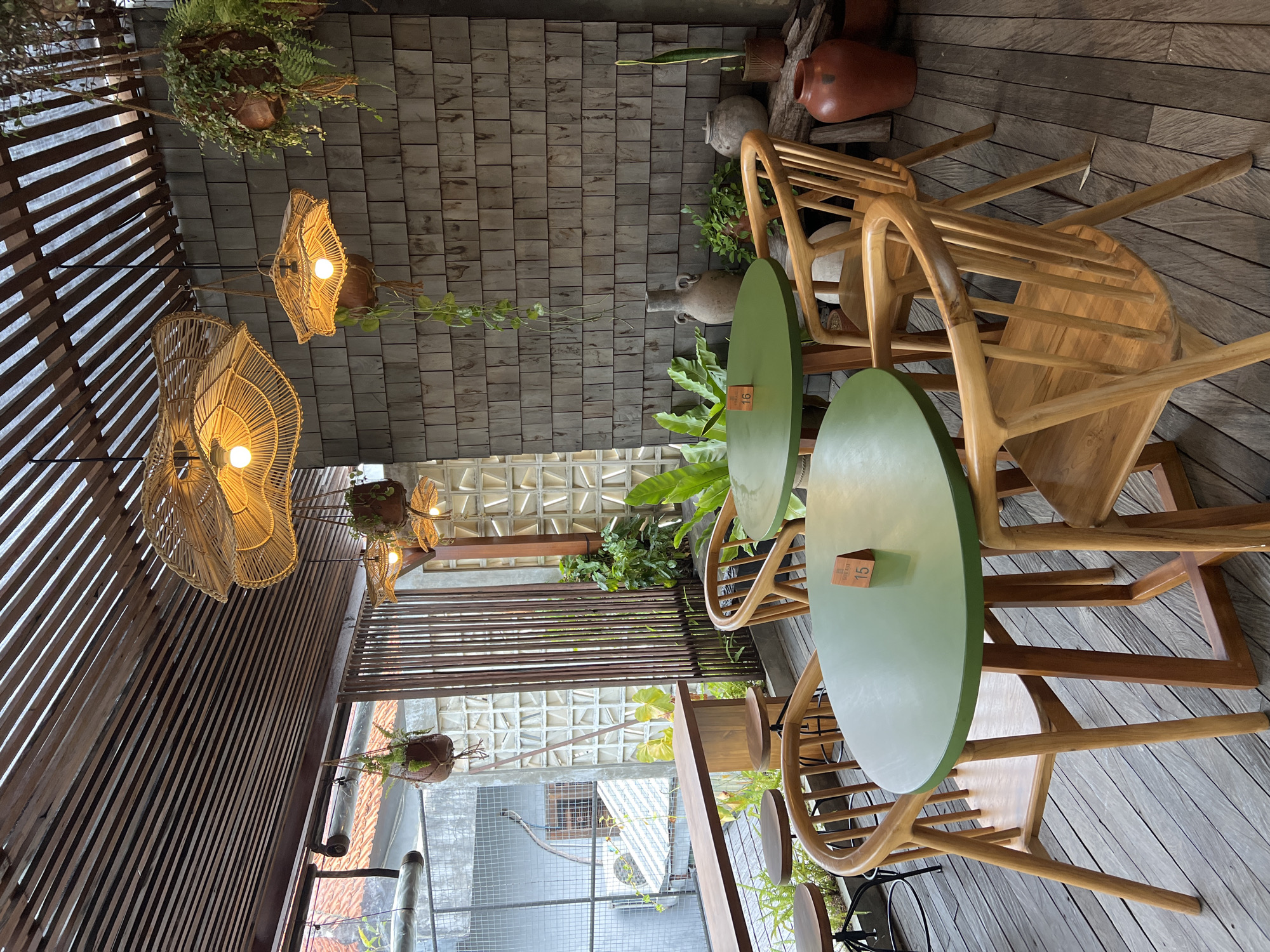 The image depicts an indoor dining area in Singakerta, Indonesia. The space is characterized by wooden flooring and a mix of ceiling treatments: some sections have a wood-shingled pattern, while others feature open wooden slats. Decorative, woven lamps hang from the ceiling, and potted plants are suspended from above.
Two round tables, painted light green, are visible, each accompanied by wooden chairs. Table number identifiers are present on the tables. Various ceramic pots and plants contribute to the decor, positioned along the edges of the dining area. A window provides a view to an exterior courtyard area.